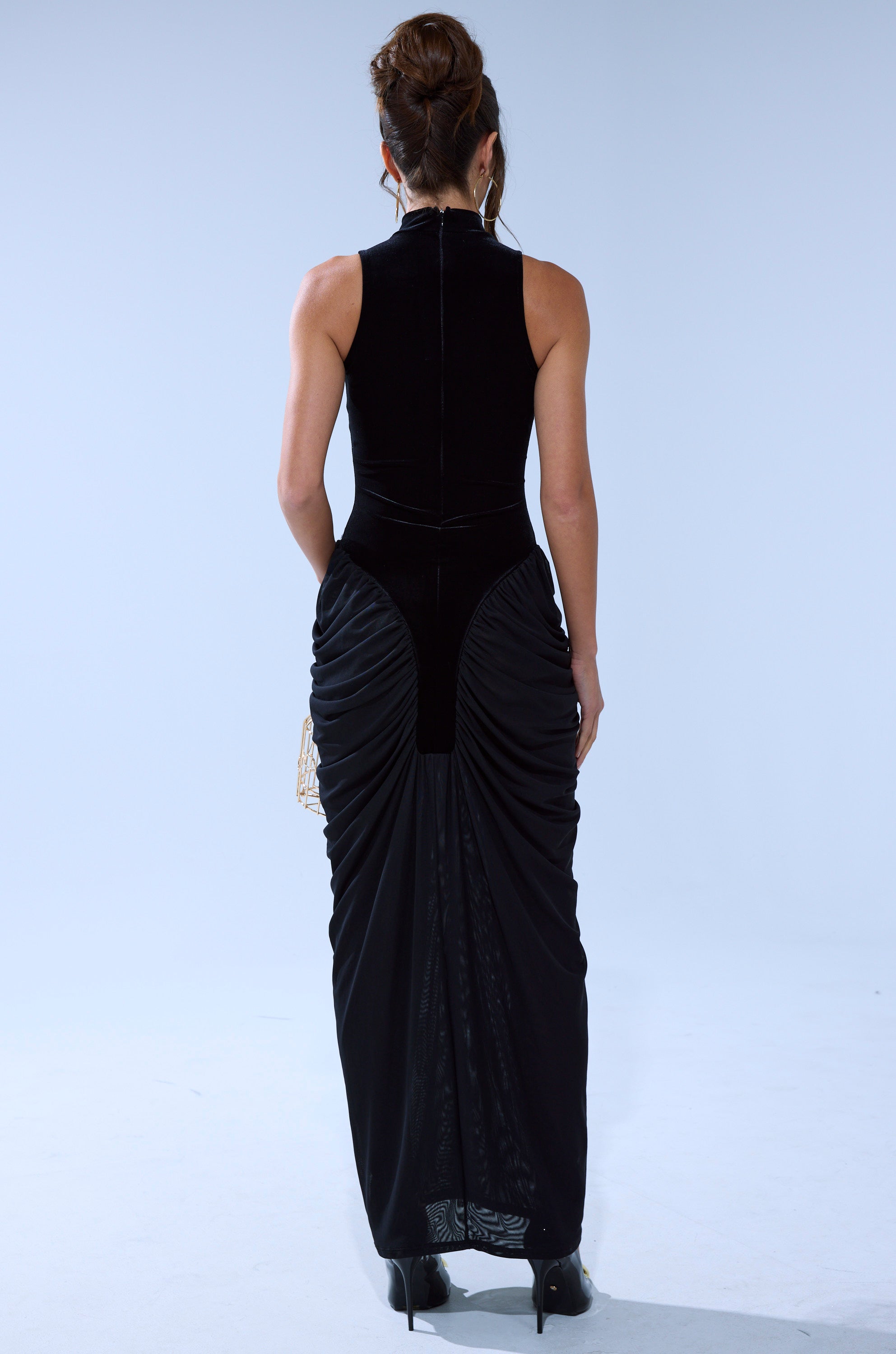 A woman with an elegant updo faces away, wearing the OUT FOR THE NIGHT MOCK NECK MAXI DRESS in black—sleeveless, with a high mock neckline and ruched back—paired with black heels against a plain light gray background.