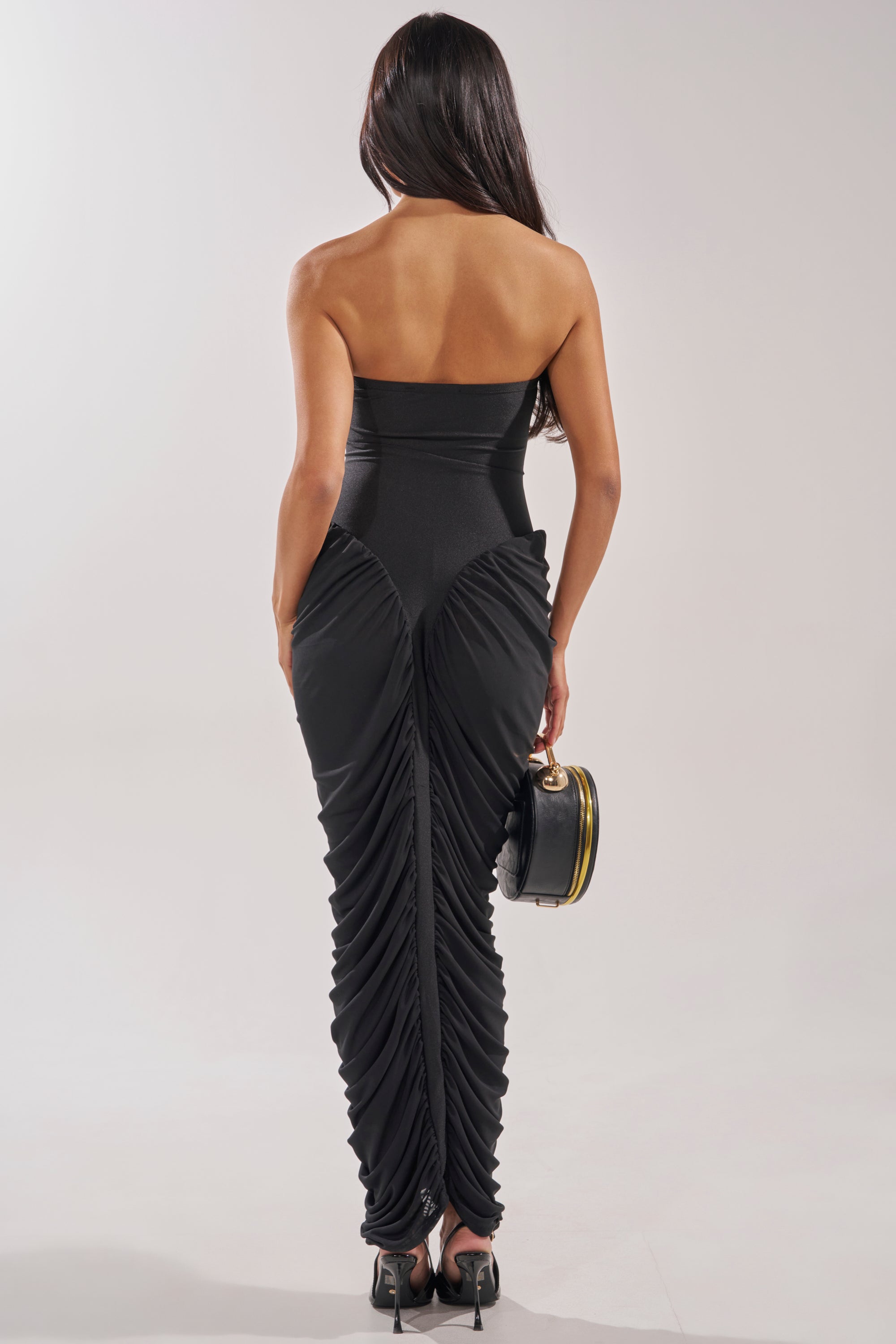 A woman with long dark hair faces away, wearing the SEASON OF SLAY STRAPLESS MAXI DRESS—a black strapless bodycon style with ruched detailing. She holds a round black handbag and pairs the look with black heels against a light background. 4