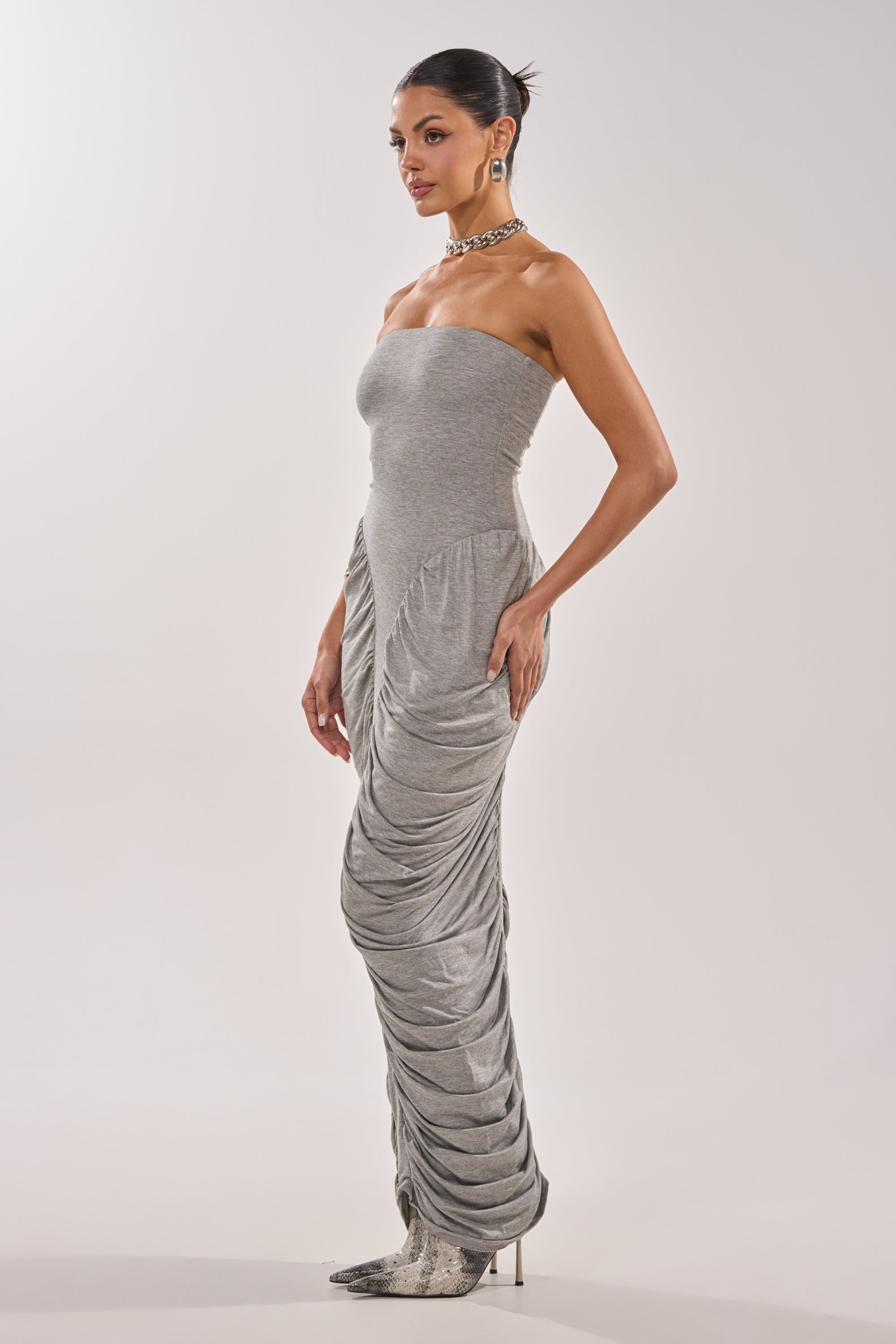 A woman in profile models the CLASSIC CHIC STRAPLESS MAXI DRESS in heather gray, featuring ruched panel detailing. She completes the look with a chunky necklace, large earrings, and pointed snakeskin heels against a plain background. 3