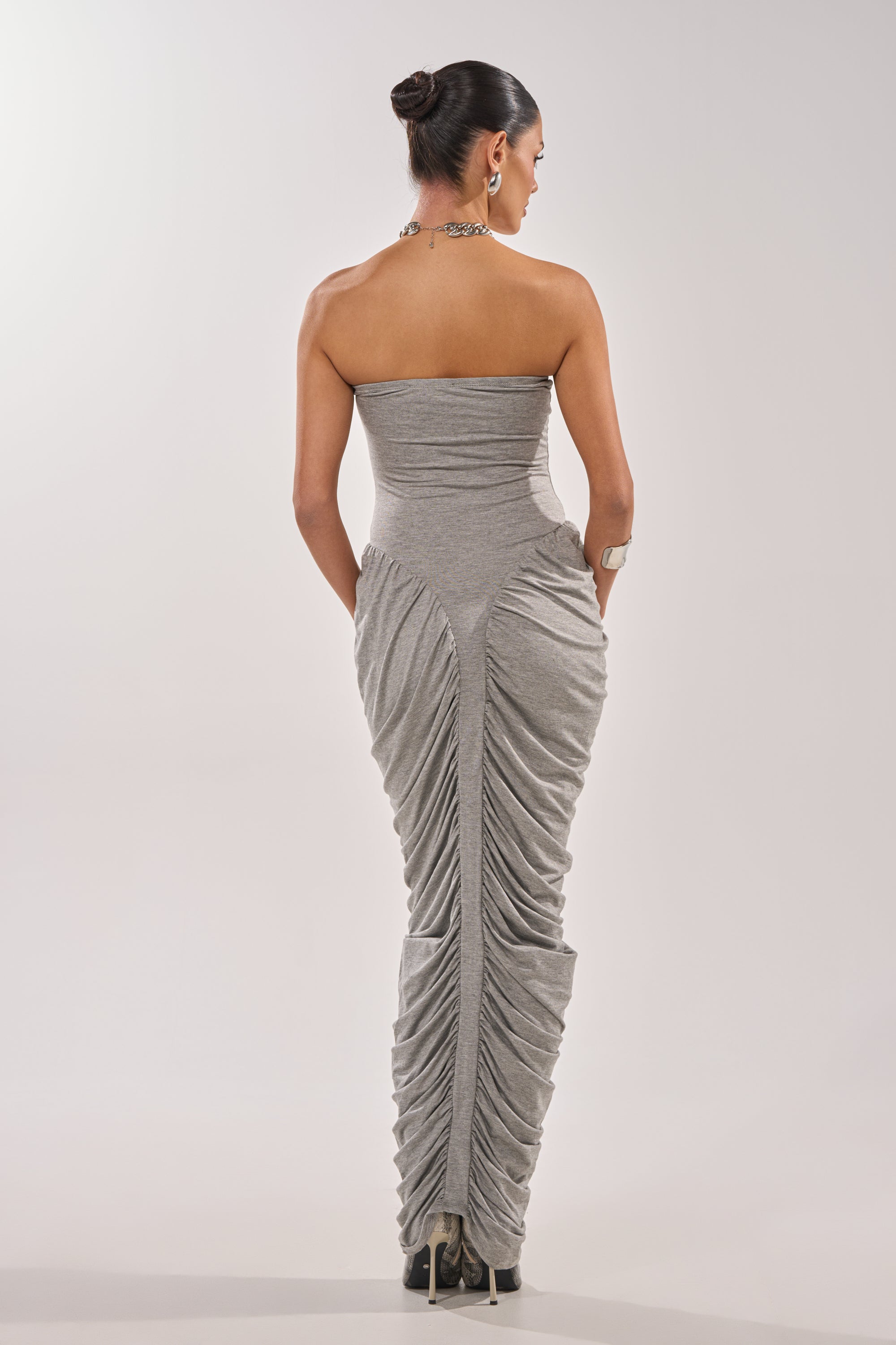 A woman with her hair in a bun is seen from behind wearing the CLASSIC CHIC STRAPLESS MAXI DRESS with ruched panels and black high-heeled boots. She accessorizes with a bracelet and necklace, standing against a plain light backdrop. 4