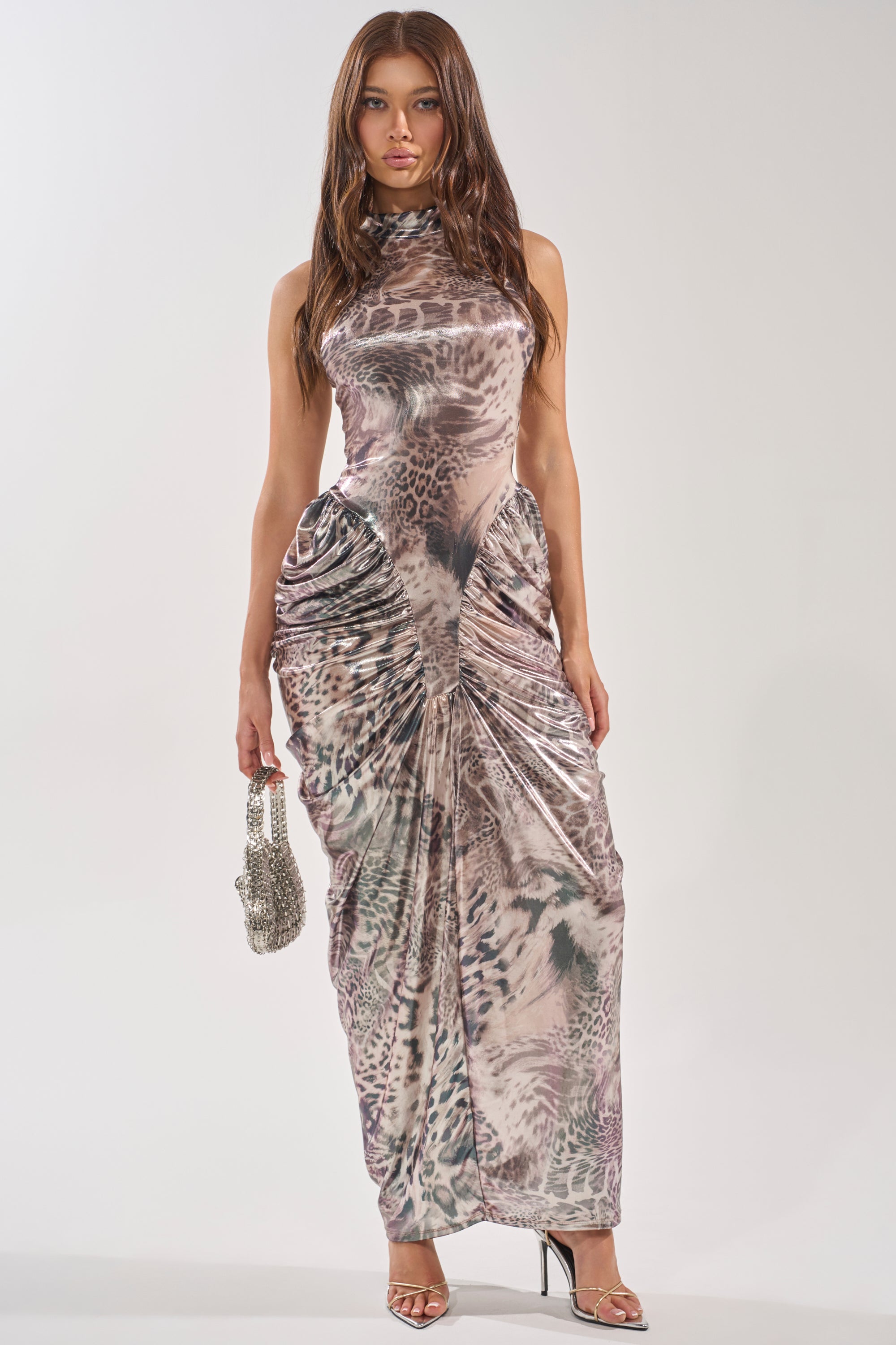 A woman stands facing forward in the STANDING MY GROUND MOCK NECK MAXI DRESS, a sleeveless, ruched maxi with an abstract animal print. She accessorizes with strappy heels and a sparkling handbag. 0