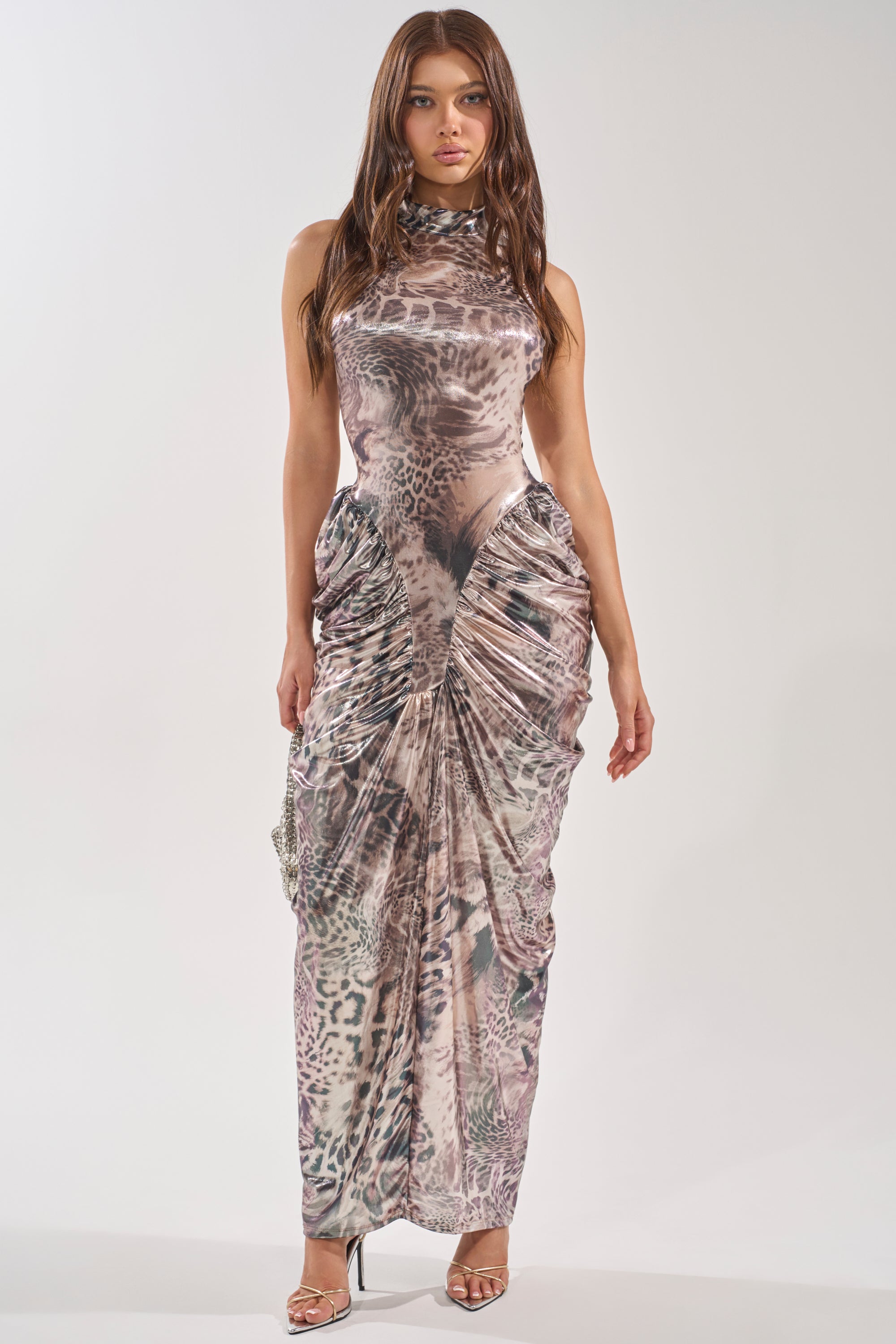 A woman faces forward in the STANDING MY GROUND MOCK NECK MAXI DRESS, a sleeveless maxi with a mock neck and ruched details. She has long brown hair and wears open-toe high-heeled sandals. 1