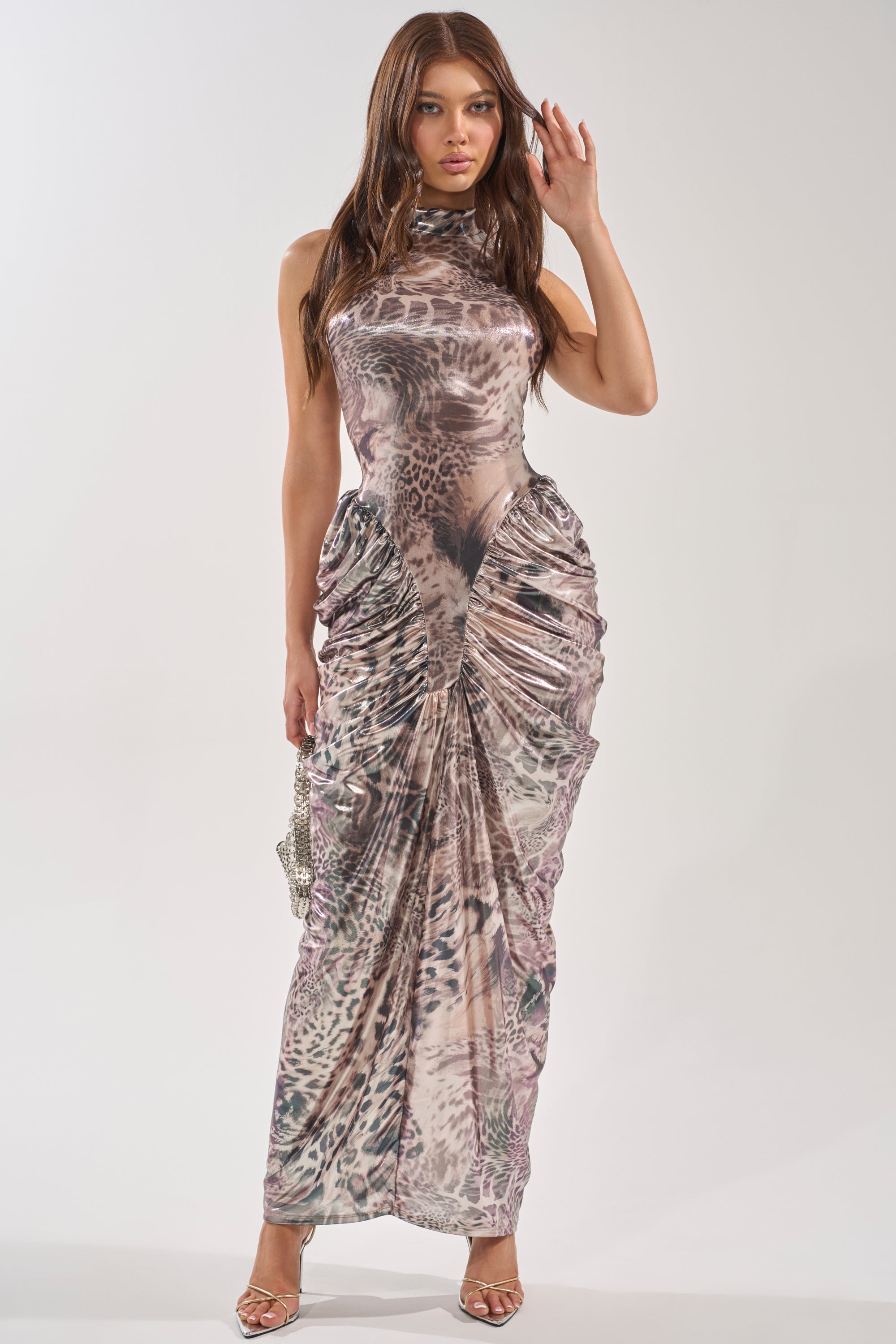 A woman stands against a plain background in the STANDING MY GROUND MOCK NECK MAXI DRESS, a fitted, sleeveless leopard print maxi with ruched detailing, accessorized with a small purse and strappy heels. 2