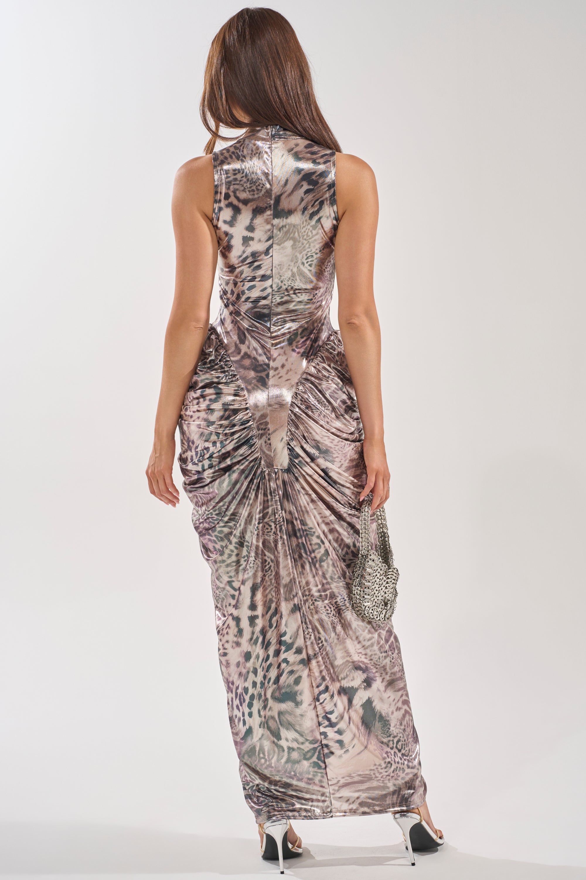 A woman with long brown hair, seen from behind, wears the STANDING MY GROUND MOCK NECK MAXI DRESS—a sleeveless, form-fitting leopard print maxi dress—paired with white heels and a silver handbag for an effortlessly chic evening look. 4