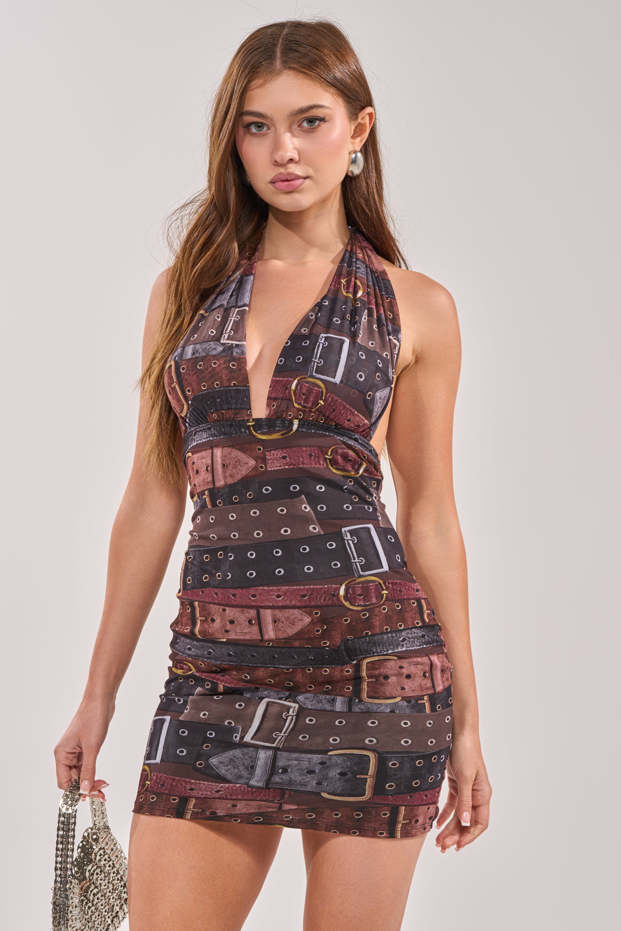 A woman with long brown hair wears the LOCKED IN PRINTED MINI DRESS IN BROWN, paired with a small silver handbag. She poses against a plain light background, highlighting her chic festival look. 1