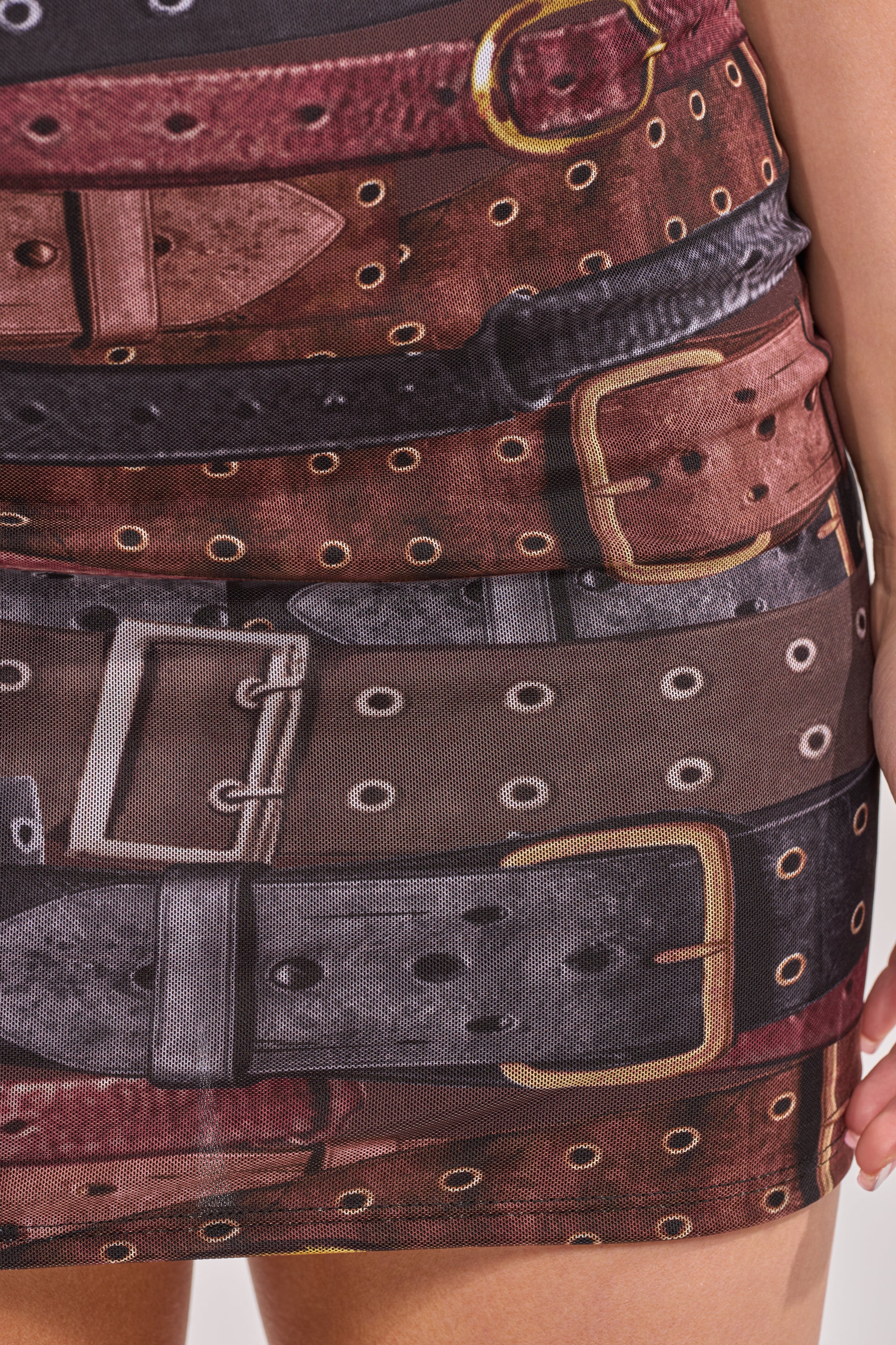 Close-up of the LOCKED IN PRINTED MINI DRESS IN BROWN, featuring overlapping brown and black belt prints with varied buckles, grommets, and stitching. A hand rests at the side—perfect for making a statement on your next night out. 8