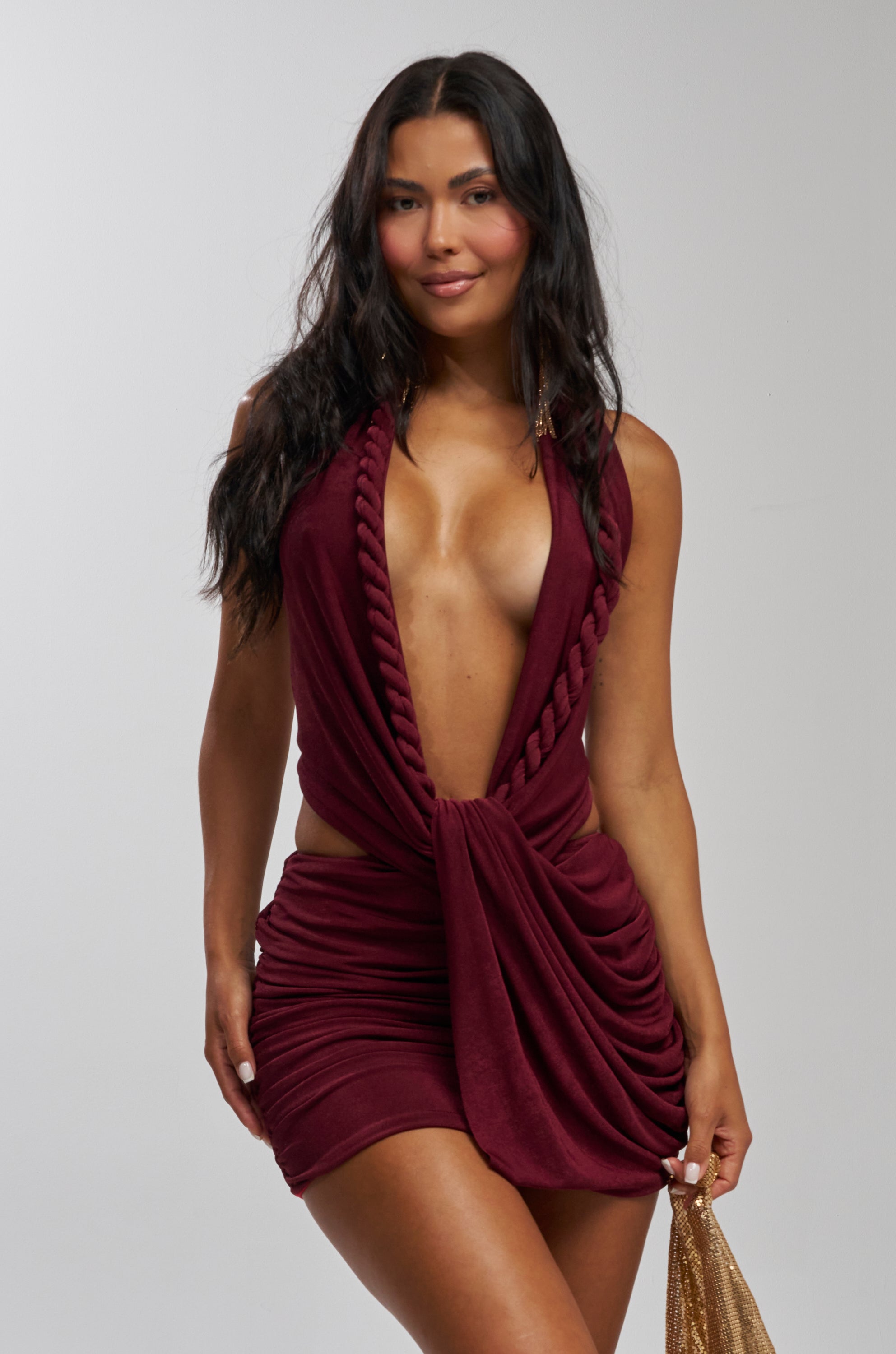 A woman with long dark hair wears the TIED TO YOU KNOTTED MINI DRESS IN BURGUNDY, featuring twisted rope details and a plunging neckline. She holds a gold handbag, smiling slightly against a plain light background—a chic vacation look.
