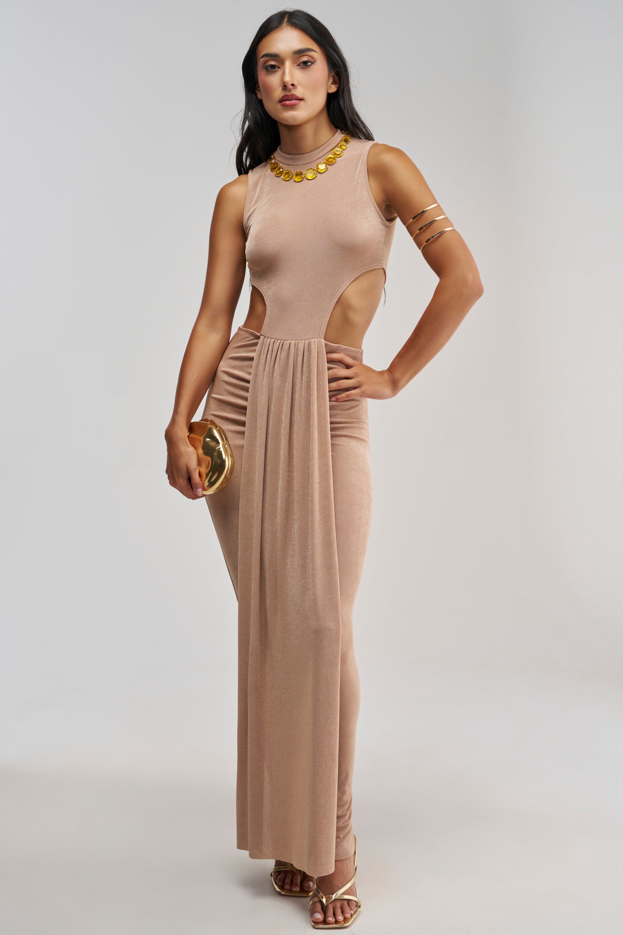 Wearing the MAKE IT UP TO ME MAXI DRESS IN CARAMEL, a woman stands against a plain light background. The sleeveless evening dress features side cutouts and a gathered drape, styled with gold accessories and heels as she holds a gold clutch.