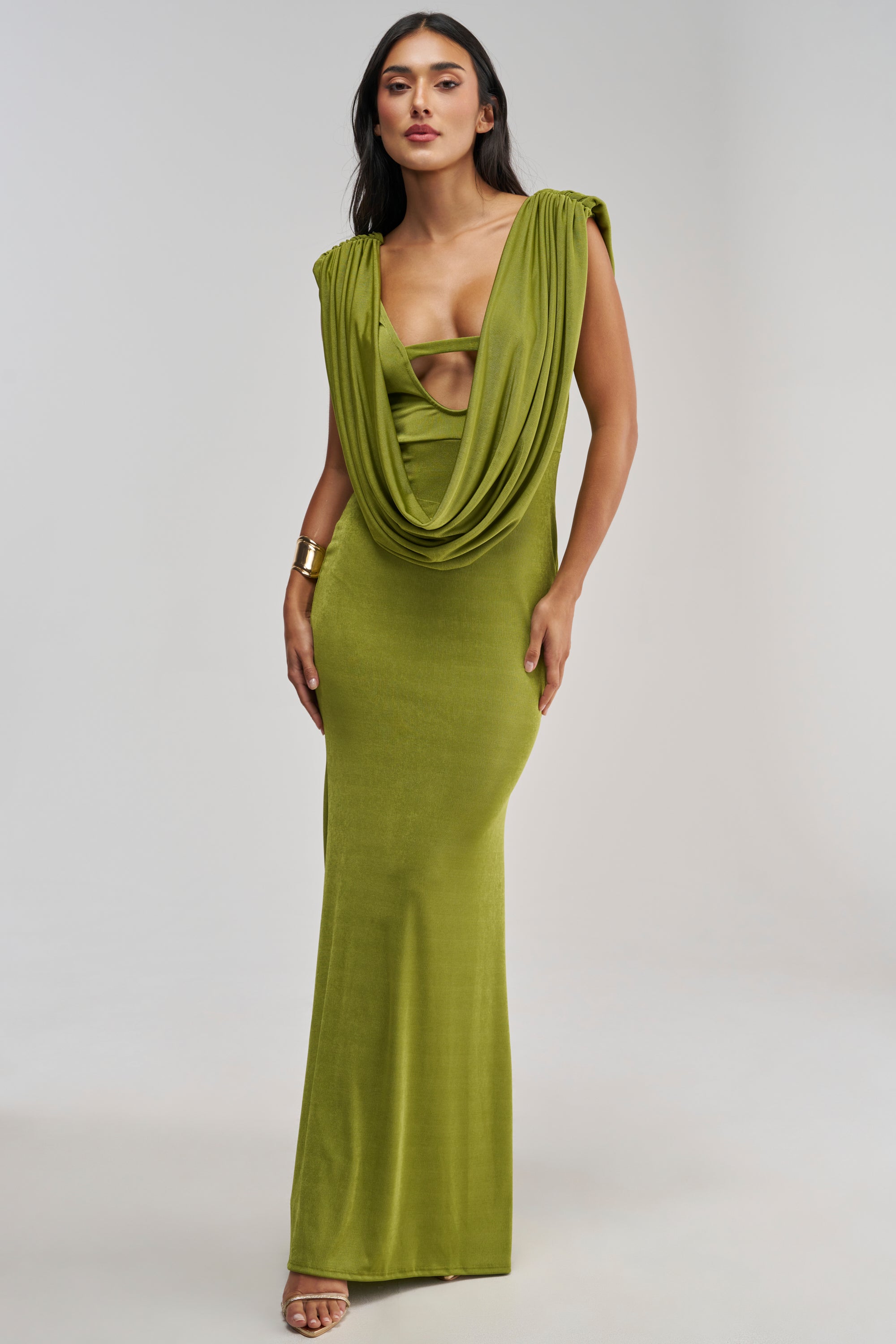 A woman stands against a plain light background wearing the HOTTER THAN EXPECTED MAXI DRESS, featuring a long olive green silhouette with draped fabric and a deep neckline, styled with gold jewelry and nude heels.