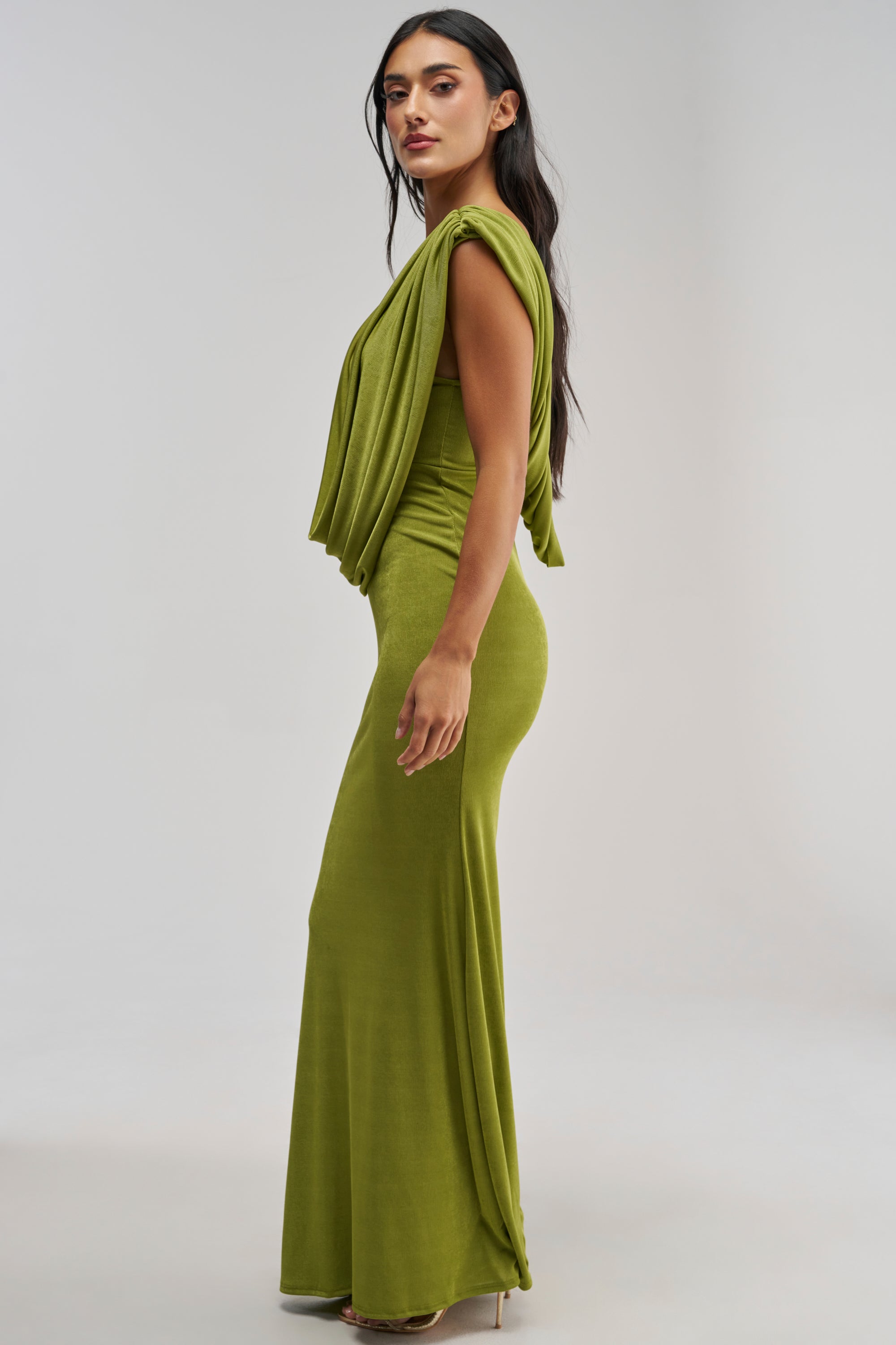 A woman stands sideways against a plain light background, looking toward the camera while wearing the HOTTER THAN EXPECTED MAXI DRESS—an olive green, long and fitted evening gown featuring draped fabric over the shoulders.