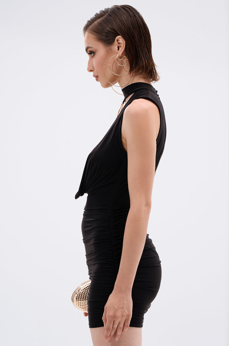 A woman with wet-look short hair and large hoop earrings stands in profile against a plain white background, wearing the CAN'T REACH ME PLUNGING MINI DRESS WITH HARDWARE and holding a small metallic handbag at her side.