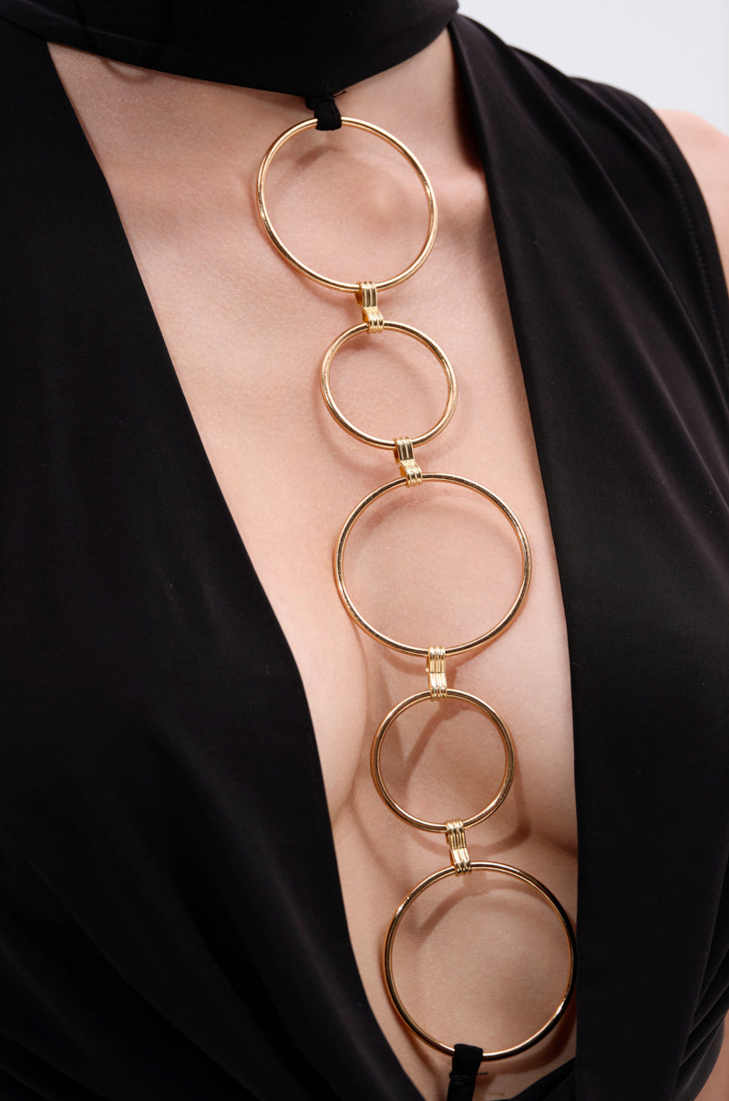 A person wearing the CAN'T REACH ME PLUNGING MINI DRESS WITH HARDWARE, a black mini dress with a deep neckline accented by bold gold hardware featuring five linked rings down the chest.