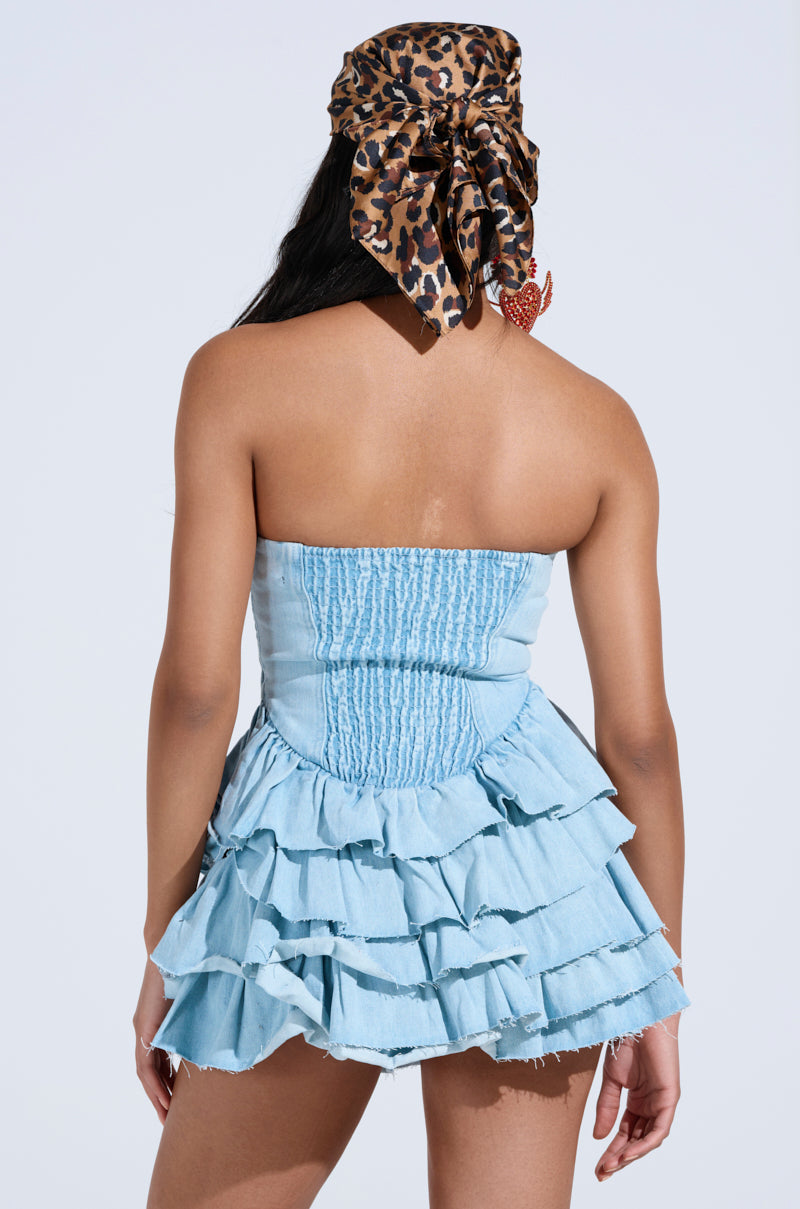 Rear view of a person wearing the AMELIA DENIM RUFFLE MINI DRESS in light blue, featuring tiered ruffles and enhanced texture, paired with a leopard print headscarf against a plain, light background.