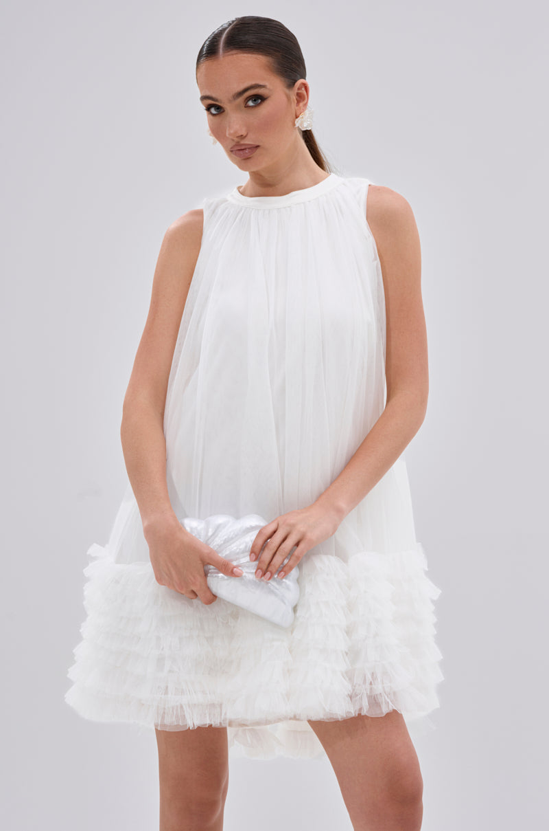 A woman wears the ARI TULLE RUFFLE MINI DRESS IN WHITE—a sleeveless, white mini dress with ruffled hem—holding a small white clutch. Her straight dark hair is pulled back as she stands against a plain light gray background.