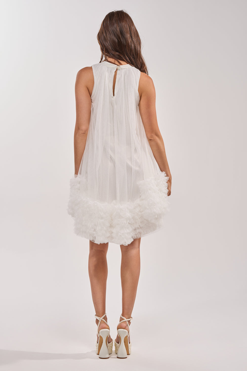 A woman stands facing away, wearing the ARI TULLE RUFFLE MINI DRESS IN WHITE—a sleeveless mini dress with sheer overlay and ruffled hem, ideal for spring evenings—paired with white strappy high heels. 4