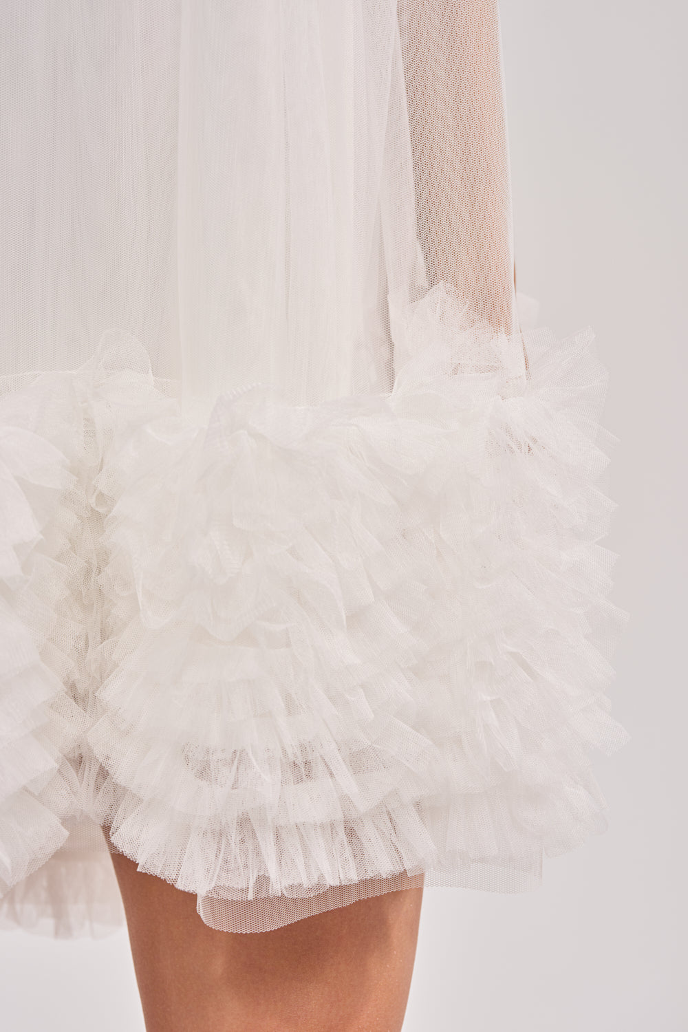 Close-up of the ARI TULLE RUFFLE MINI DRESS IN WHITE, showcasing layered white tulle ruffles and a hint of bare leg against a neutral background. 5