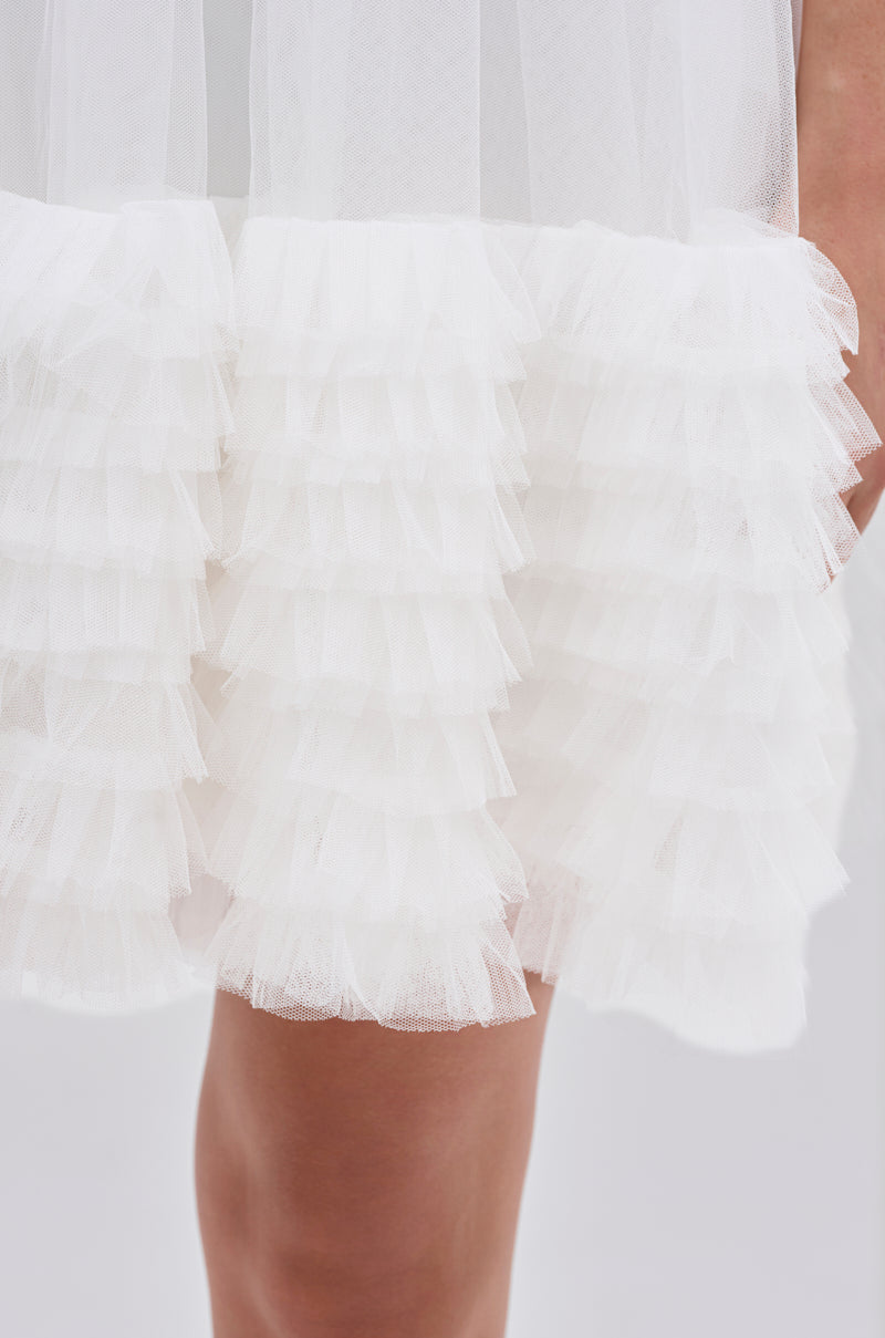 A close-up of the ARI TULLE RUFFLE MINI DRESS IN WHITE features multiple ruffled layers and accordion-pleated tulle, with the dress hem falling above a person's bare leg.