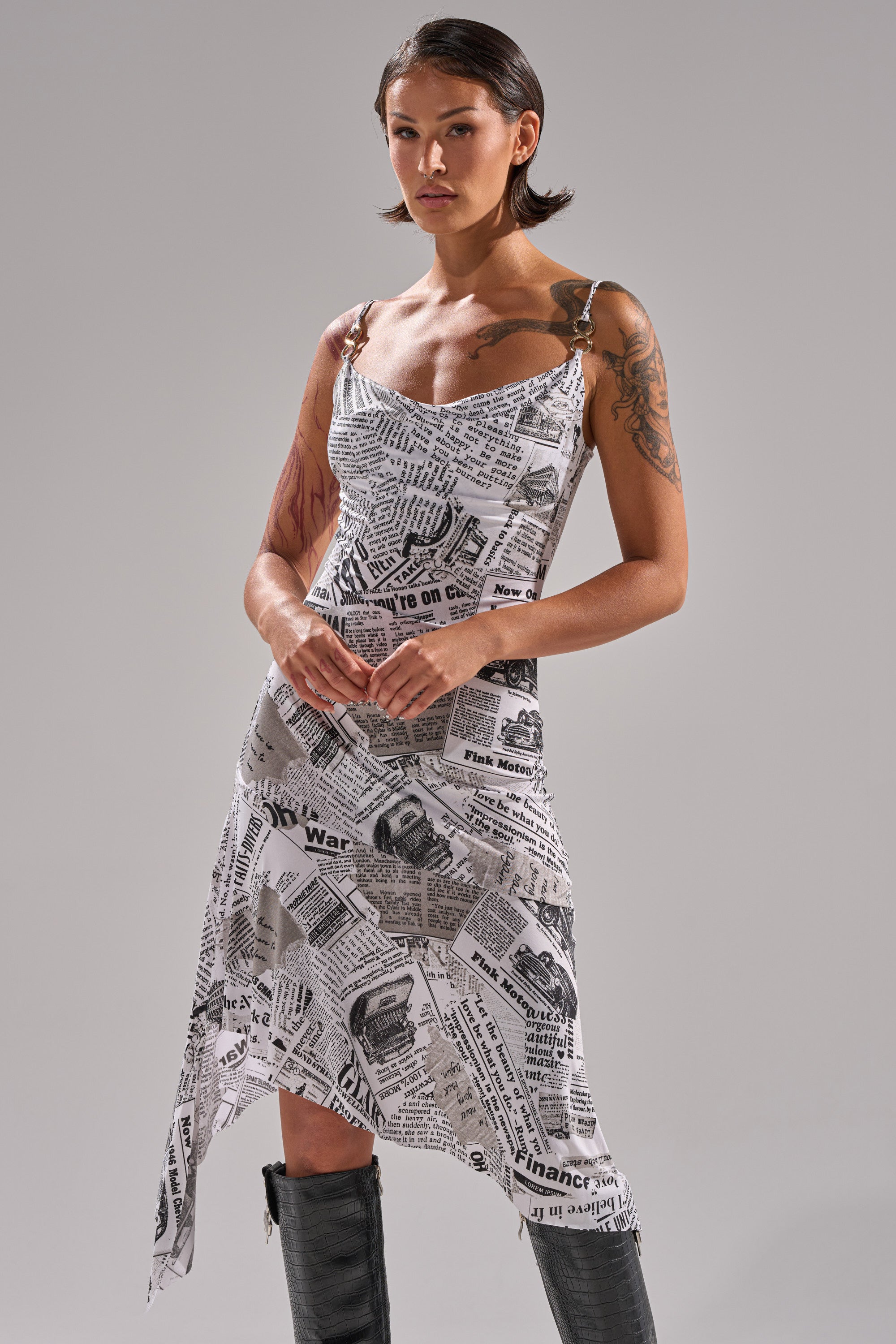 A woman with short hair and visible arm tattoos models the BREAKING NEWS PRINTED MIDI DRESS, featuring a curve-hugging black-and-white newspaper print, against a plain background. She pairs it with tall black boots.