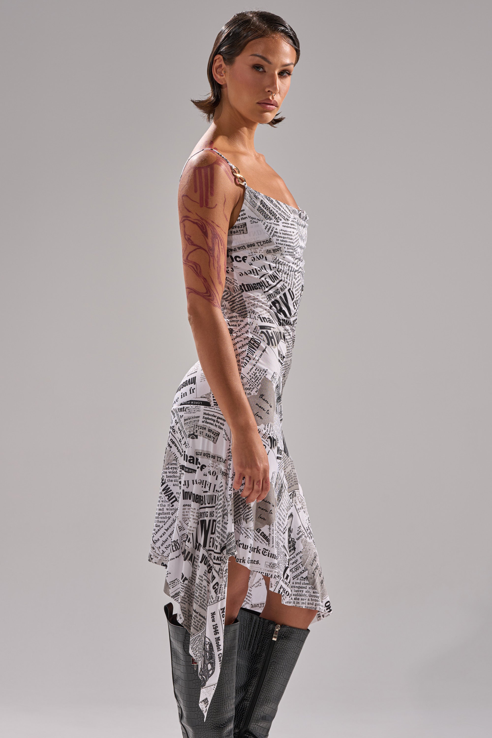 A woman in profile wears the BREAKING NEWS PRINTED MIDI DRESS—an asymmetrical white newspaper print piece—paired with black knee-high boots and bold red body paint on her right arm, set against a plain gray background.