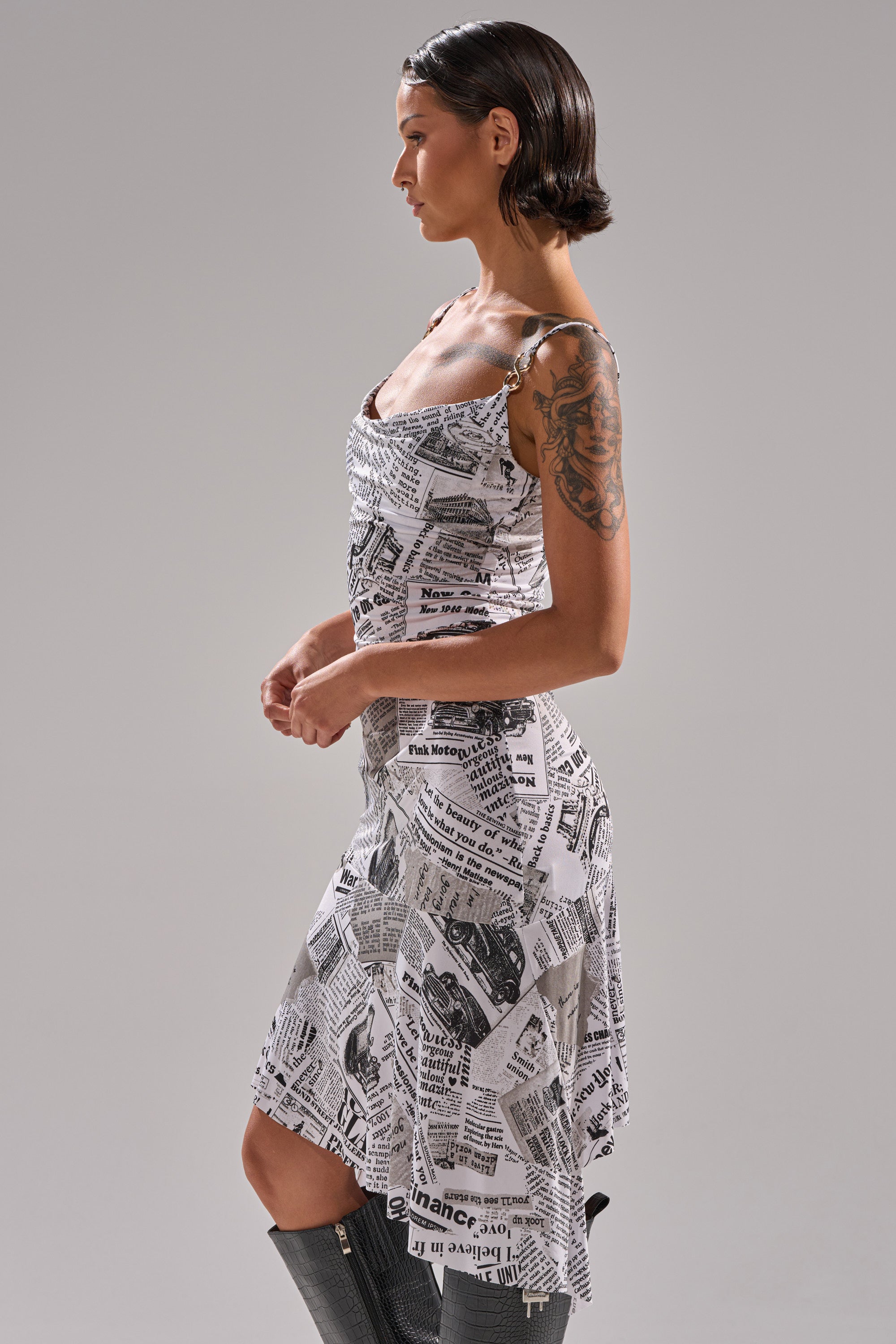 A woman with short dark hair and a visible arm tattoo models the BREAKING NEWS PRINTED MIDI DRESS, featuring black-and-white newspaper text and images, as she stands in profile against a plain gray background.
