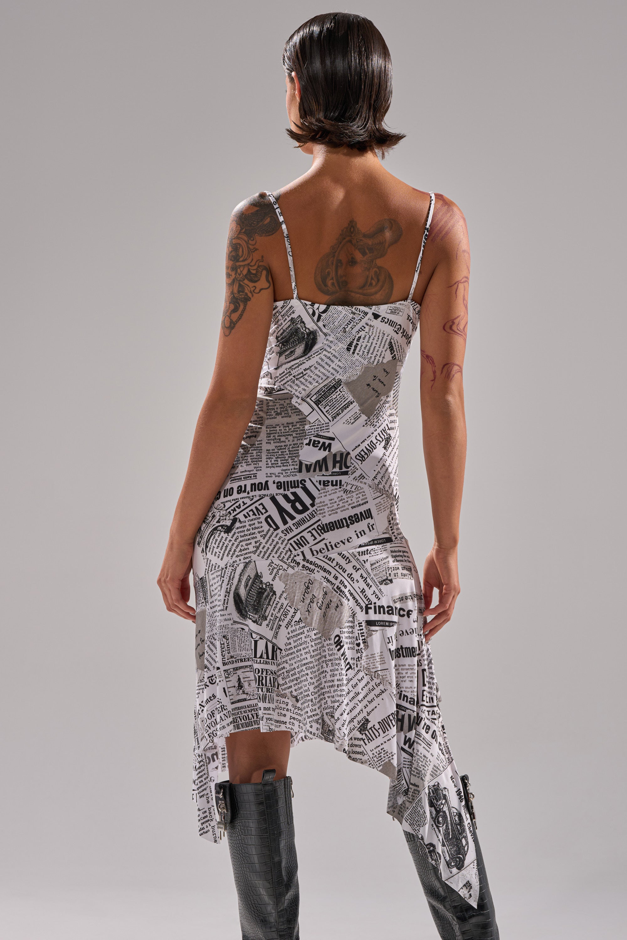 A person with short dark hair faces away, wearing the BREAKING NEWS PRINTED MIDI DRESS with an asymmetrical hem and tall black boots. The newspaper print of the dress pops against a plain gray background.