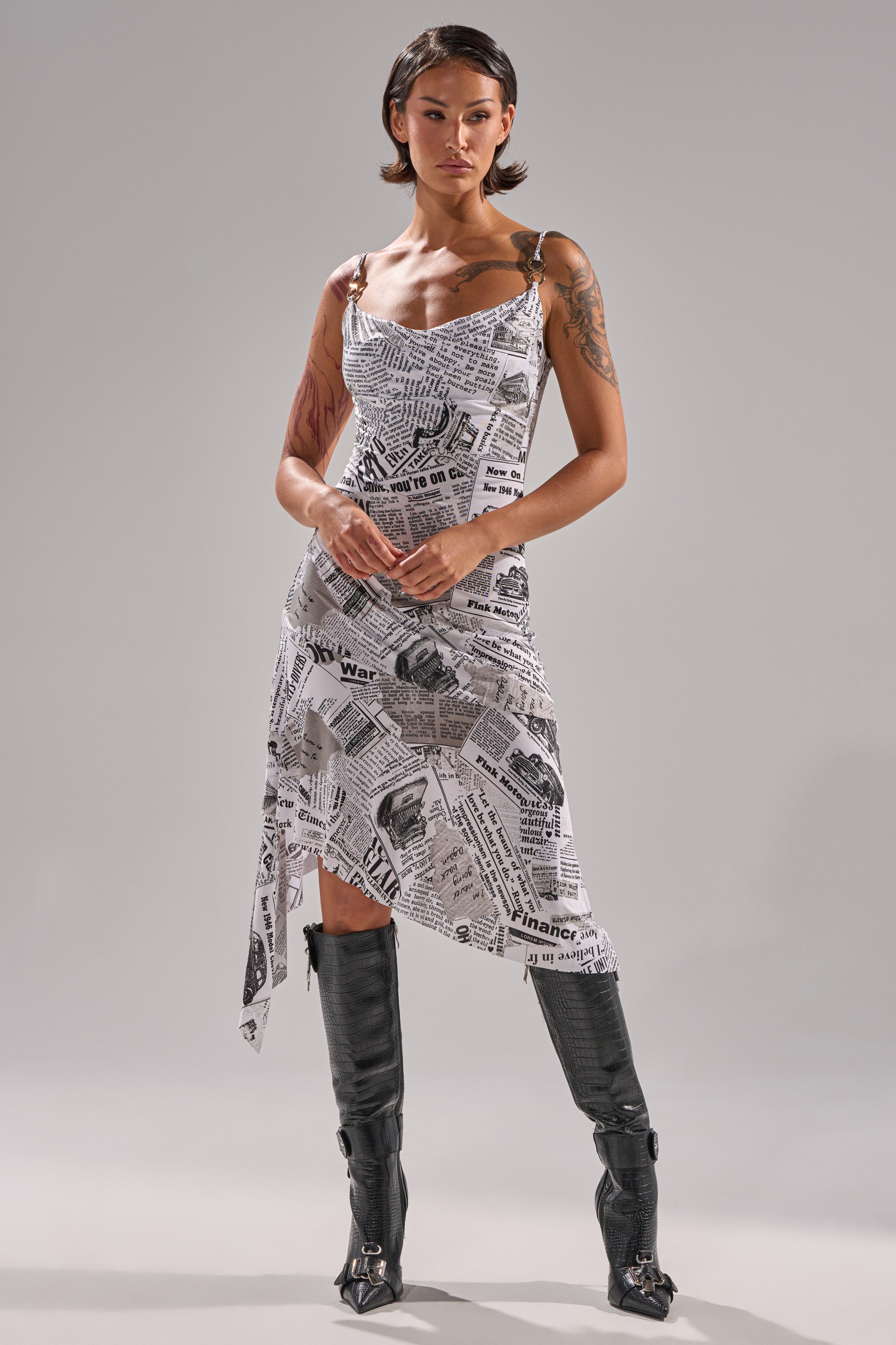 A woman with short hair and visible arm tattoos poses confidently against a gray background, wearing the BREAKING NEWS PRINTED MIDI DRESS—curve-hugging and sleeveless—paired with tall black boots.