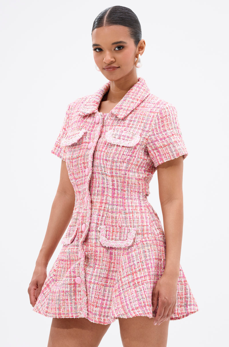 A woman poses confidently against a plain white background wearing the TAKE NOTES TWEED MINI DRESS IN PINK, featuring a pink and white plaid pattern, structured short sleeves, flared skirt, front buttons, and decorative pockets.