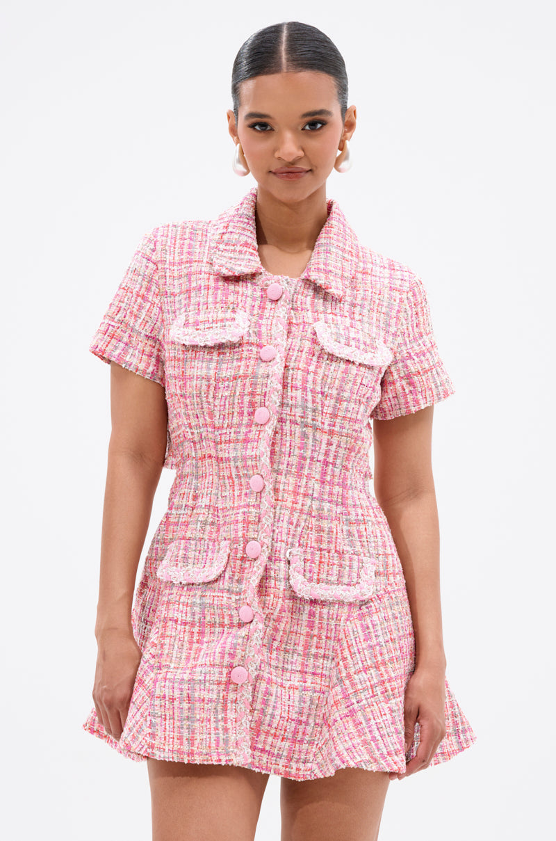 A woman stands facing forward against a plain white background, wearing the TAKE NOTES TWEED MINI DRESS IN PINK with structured short sleeves, four front pockets, and large buttons.