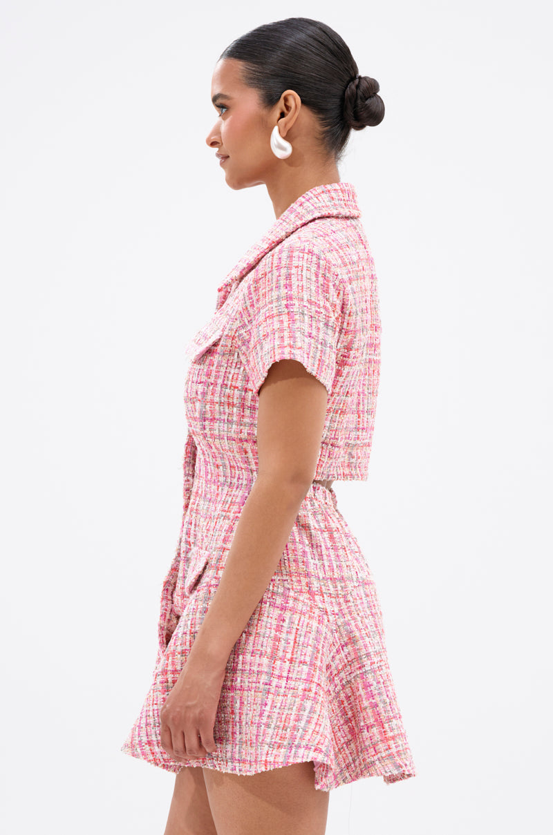 A woman with her hair in a low bun models the TAKE NOTES TWEED MINI DRESS IN PINK, which features structured short sleeves and a belted waist, as she stands in profile against a plain white background.