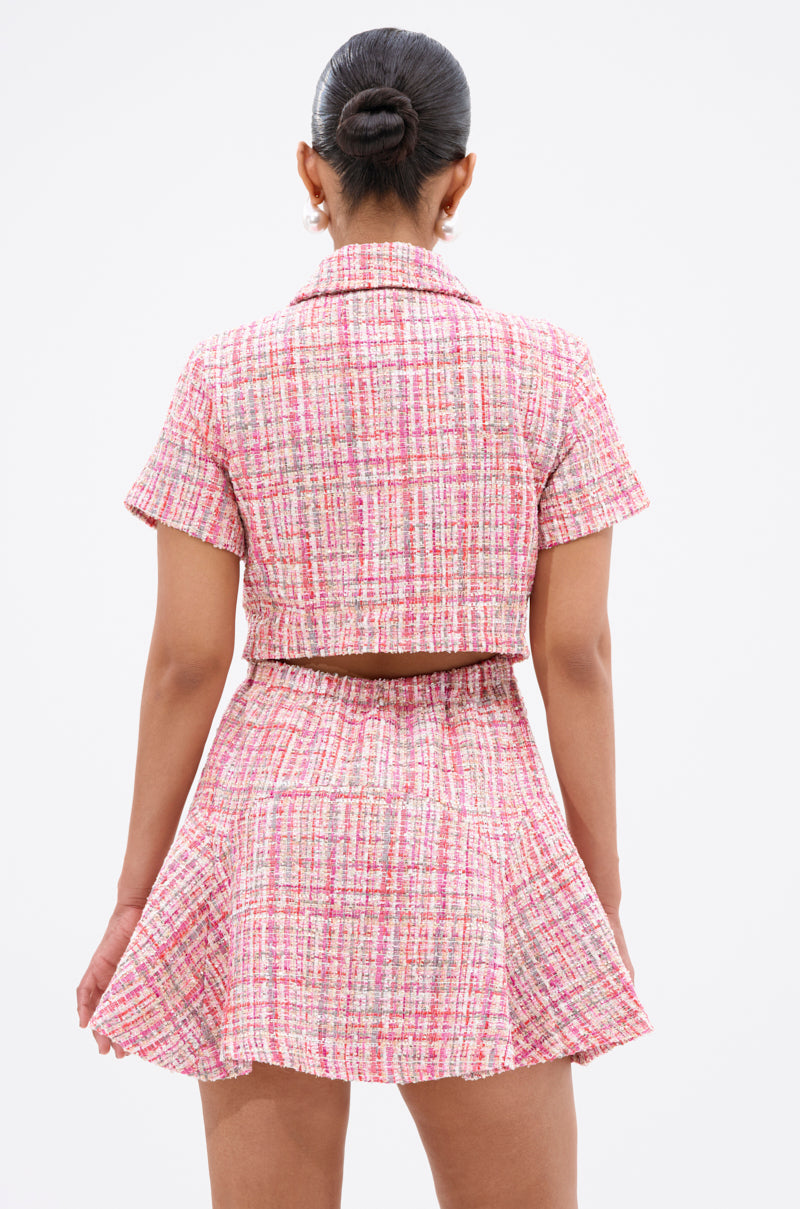 A person with dark hair in a bun models the TAKE NOTES TWEED MINI DRESS IN PINK, featuring short sleeves, a flared skirt, and high-waisted silhouette. The outfit is shown from the back against a plain white background.