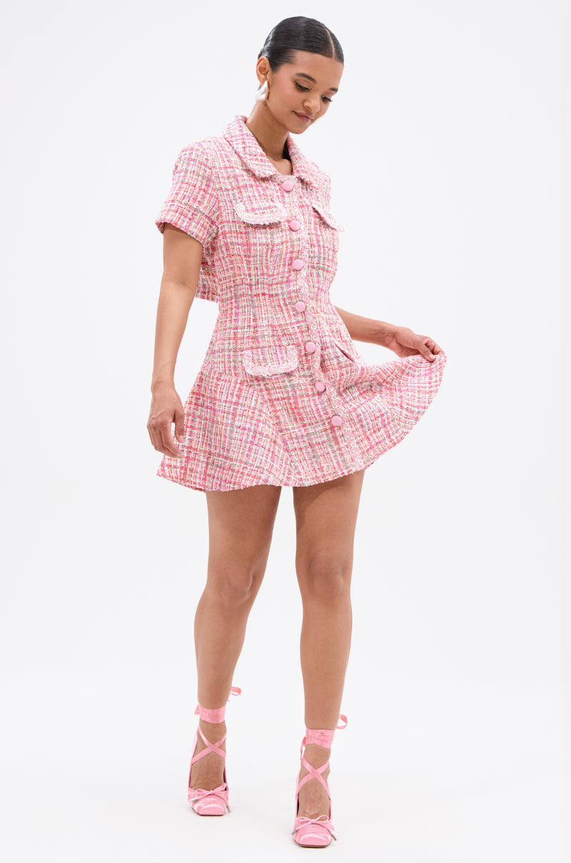 A woman smiles while holding the hem of her TAKE NOTES TWEED MINI DRESS IN PINK, a structured mini dress with short sleeves and large pockets, styled with matching pink high-heeled shoes against a white background.