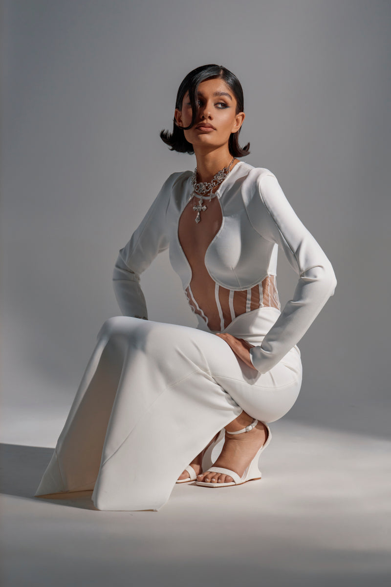 A woman with short dark hair kneels confidently, modeling the A NIGHT OUT BANDAGE MAXI DRESS IN WHITE with sheer mesh paneling and accessorizing with white sandals and a statement necklace against a softly lit background.