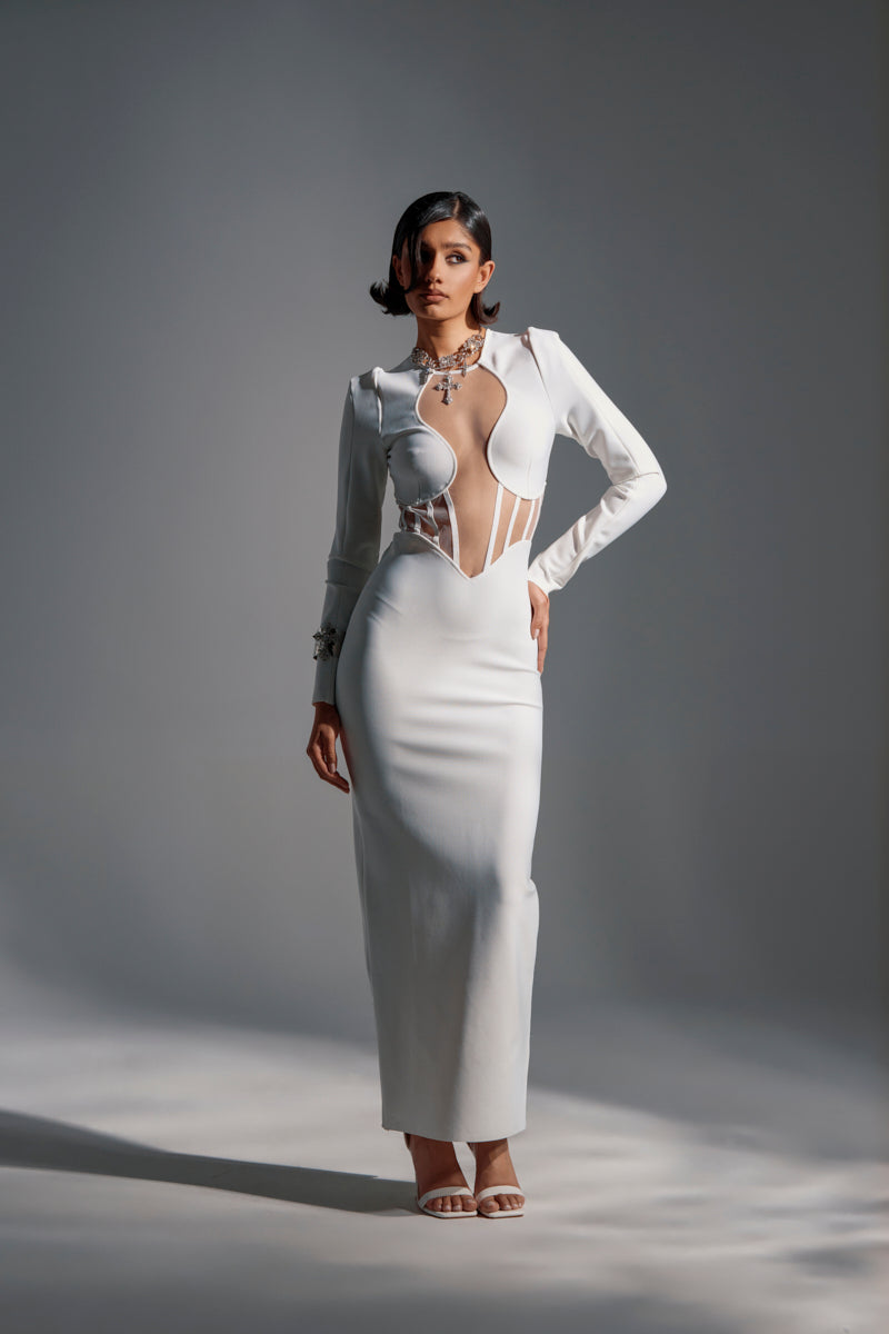 A woman poses confidently in the A NIGHT OUT BANDAGE MAXI DRESS IN WHITE, featuring a sculpted bust and cut-out accents. She accessorizes with statement jewelry and strappy heels, standing against a plain studio background.