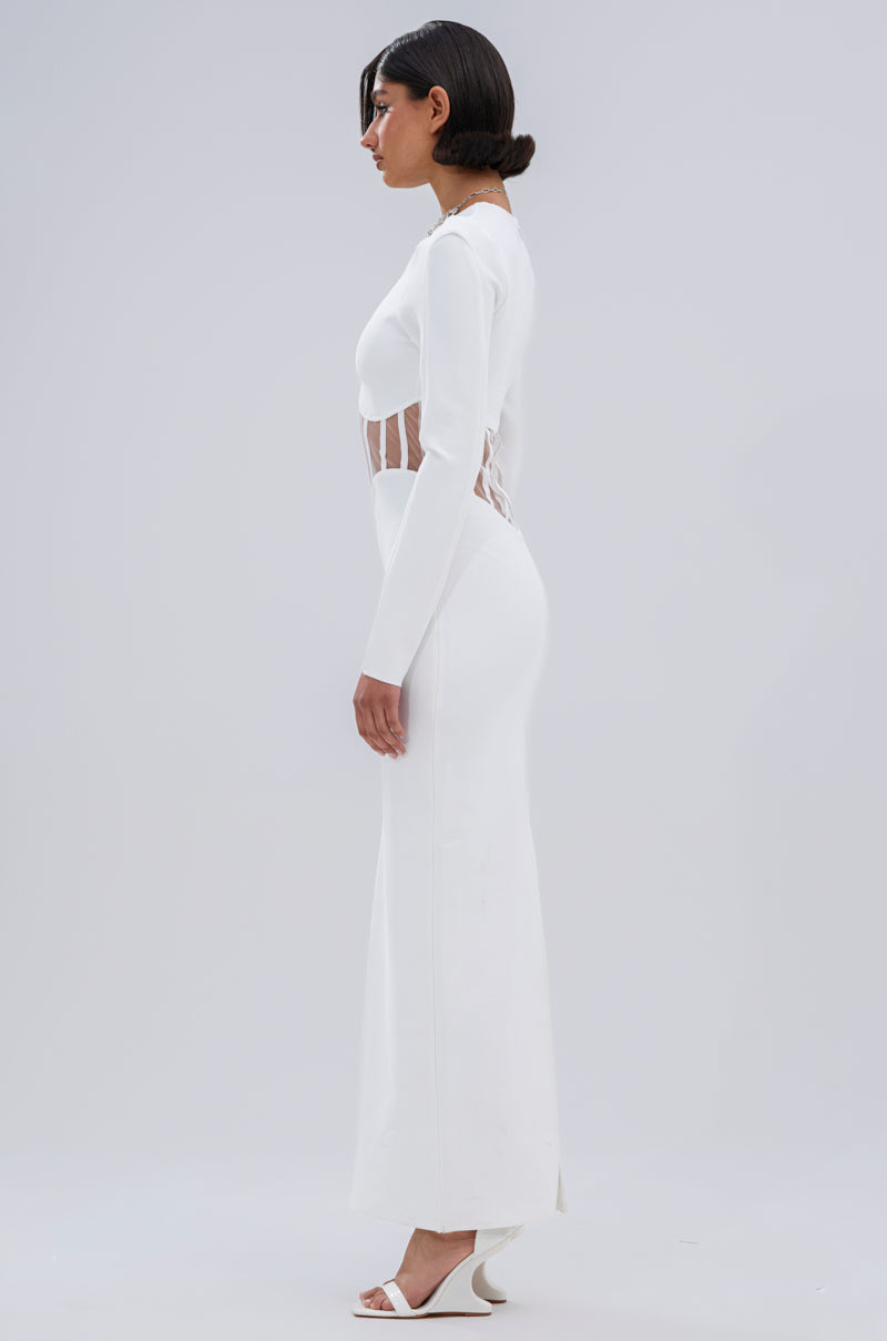 A woman in the A NIGHT OUT BANDAGE MAXI DRESS IN WHITE, featuring sheer mesh paneling, stands in profile against a light gray background, wearing white heeled sandals with her hair in a sleek low bun.