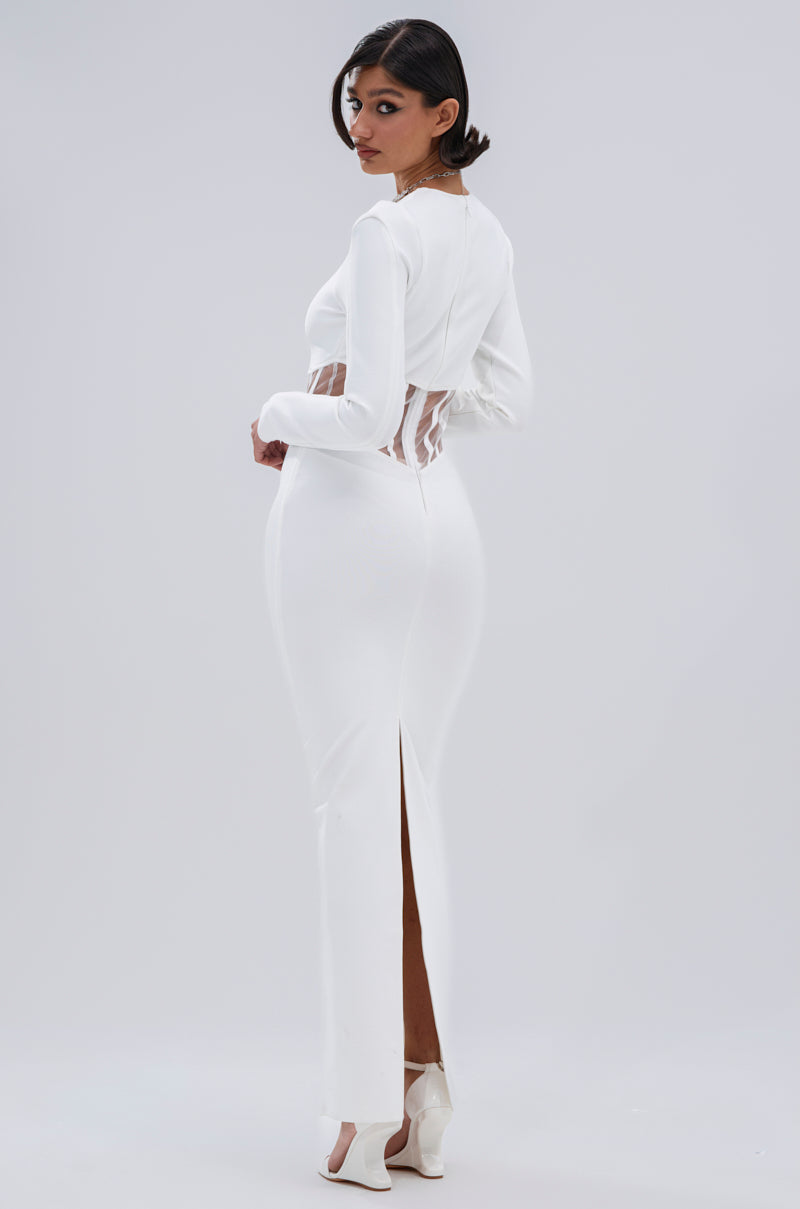 In the A NIGHT OUT BANDAGE MAXI DRESS IN WHITE featuring long sleeves, a fitted silhouette, sheer mesh panels, and a back slit, a woman stands slightly turned to the side, glancing over her shoulder against a simple light backdrop.