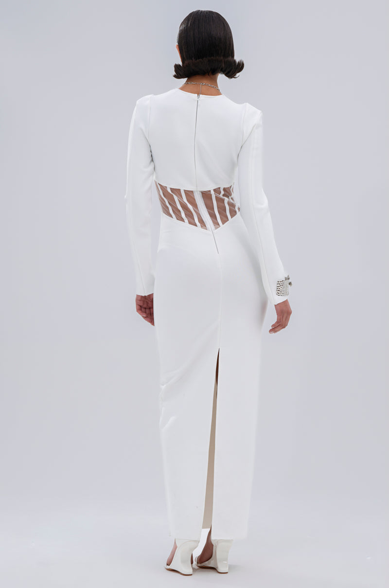 A person wears the A NIGHT OUT BANDAGE MAXI DRESS IN WHITE, a long-sleeved, floor-length gown featuring sheer mesh paneling at the back and a high back slit, highlighting its structured design and sleek hairstyle as they face away.