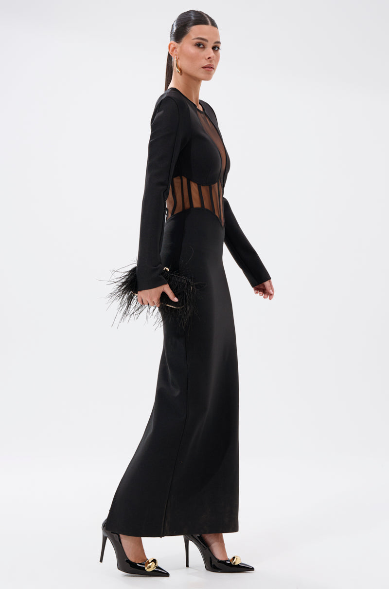 A woman walks in the A NIGHT OUT BANDAGE MAXI DRESS, a curve-hugging black maxi with sheer panels. She carries a black feathered clutch, wears black heels with gold accents, and has her hair in a sleek ponytail against a plain white background.