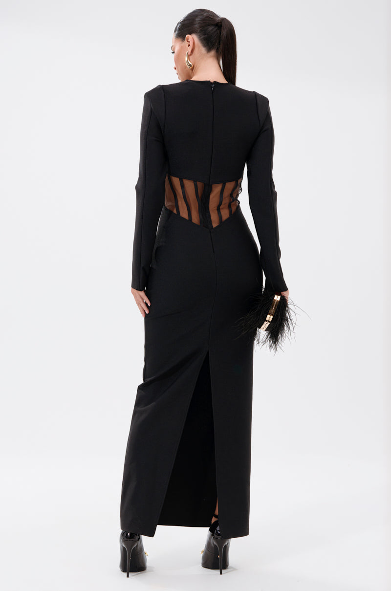 A woman in the A NIGHT OUT BANDAGE MAXI DRESS, featuring sheer mesh waist panels, long sleeves, and a back slit, stands facing away with a ponytail and holds a feather-detailed black clutch.