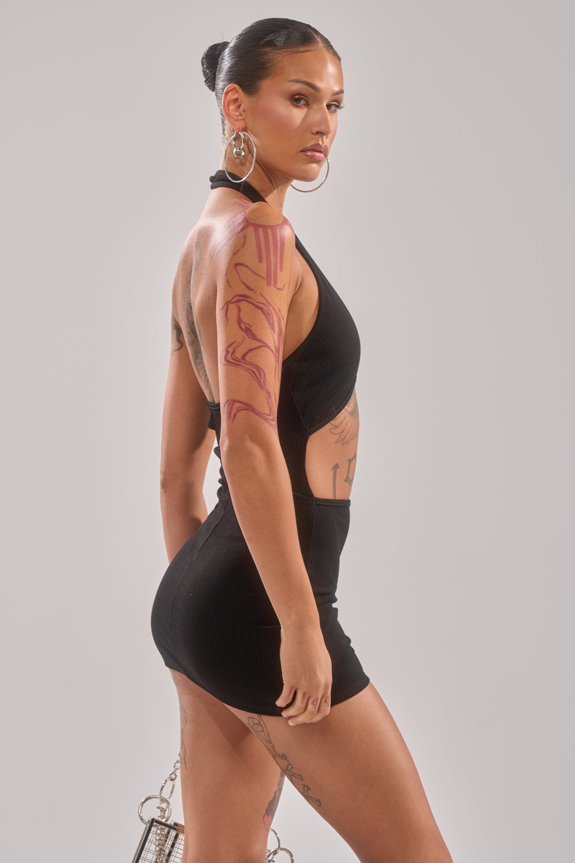 A person with long brown hair in a sleek bun, bold tattoos, and large hoop earrings confidently models the HOT AF MINI DRESS—a black sleeveless mini dress—against a plain background.