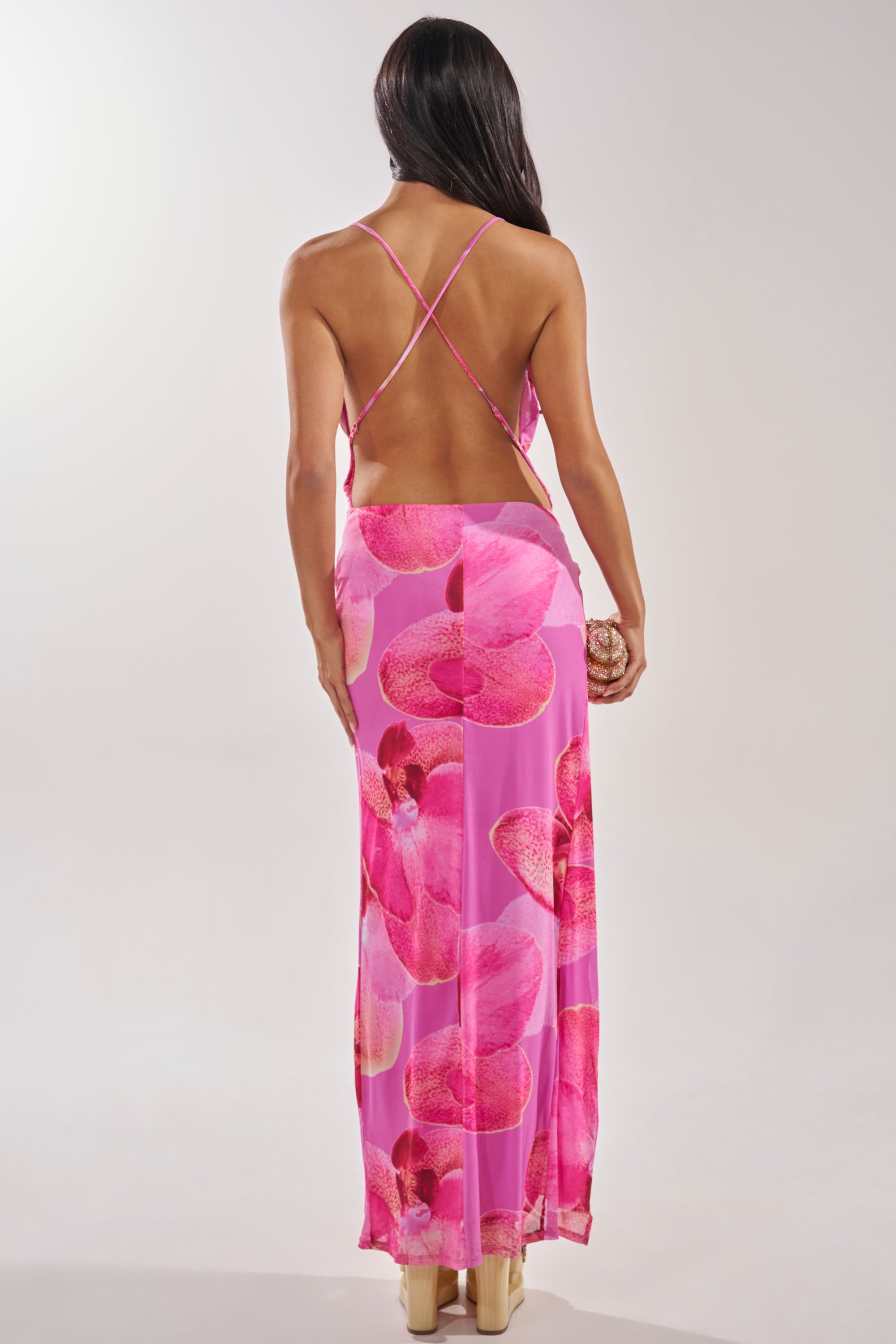 A woman with long dark hair faces away, wearing the FOREVER HER FLORAL MAXI DRESS featuring large pink patterns and crisscross straps, paired with beige heels and a small clutch, against a light background. 3