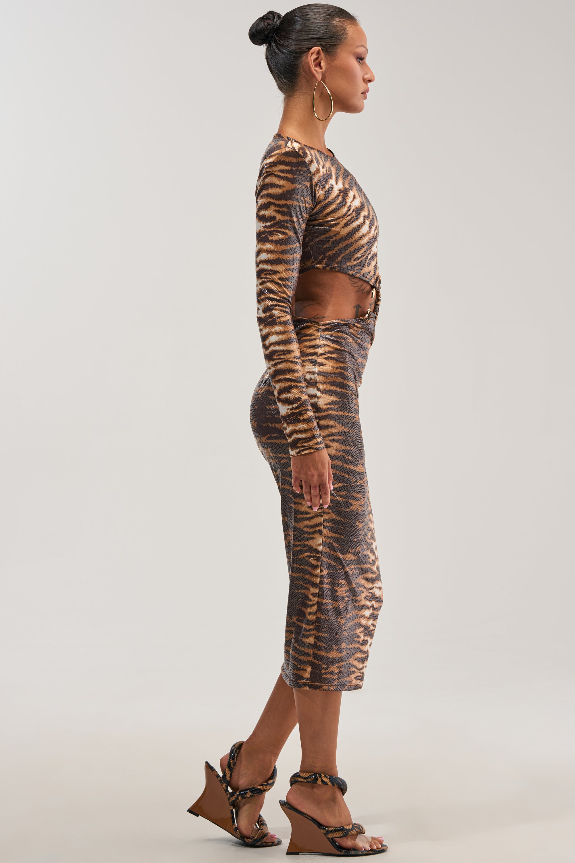 A woman in profile wears the MIXED SIGNALS ANIMAL PRINT MIDI DRESS with long sleeves, styled with a brown belt, large hoop earrings, and wedge sandals. Her hair is in a bun, and she poses against a plain light background.