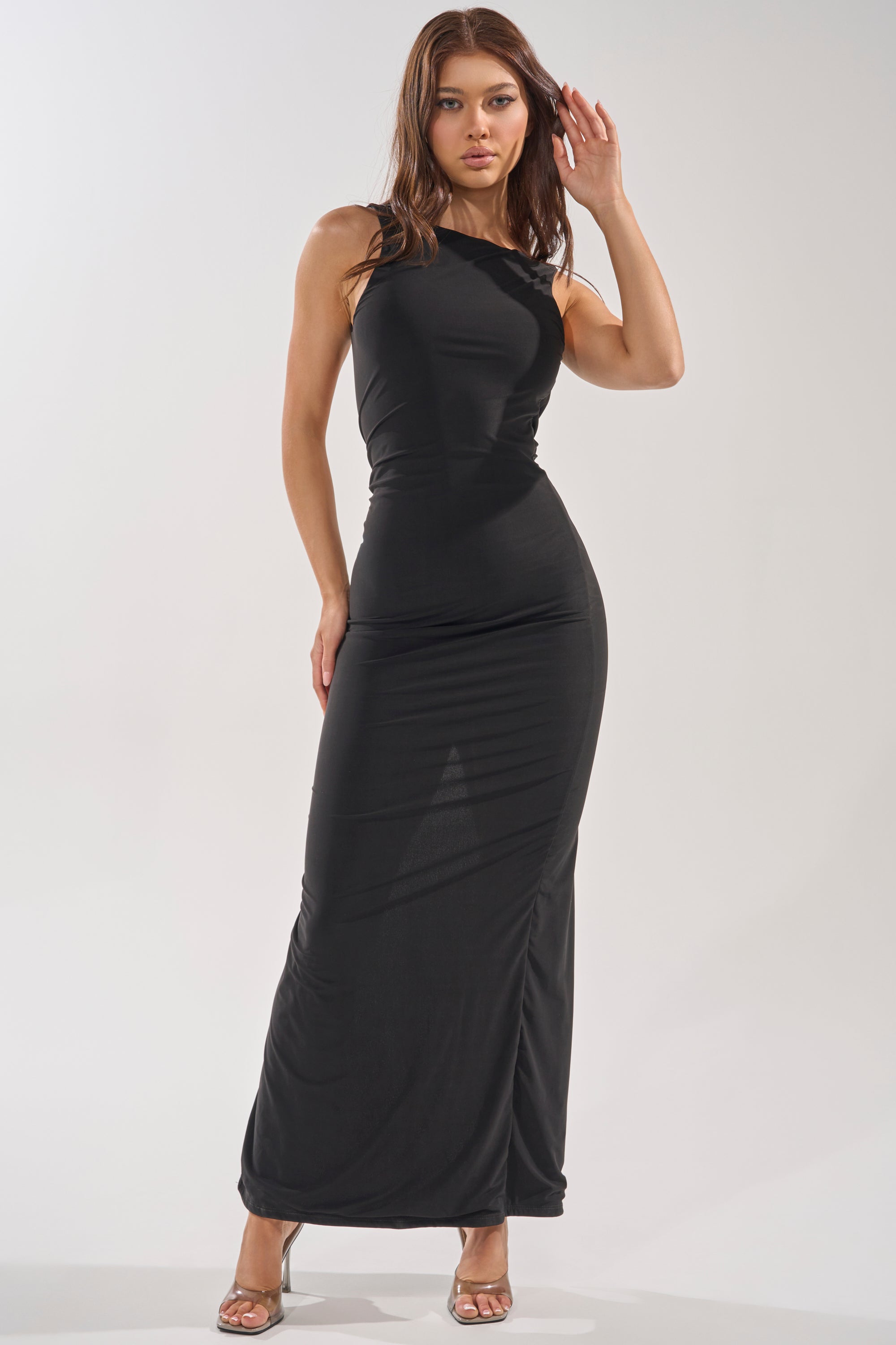 A woman poses in the ON VACA MODE LOW BACK MAXI DRESS, a sleeveless, fitted black maxi dress featuring a low-cut back, styled with silver high-heeled sandals for an elegant evening look against a simple light background. 2