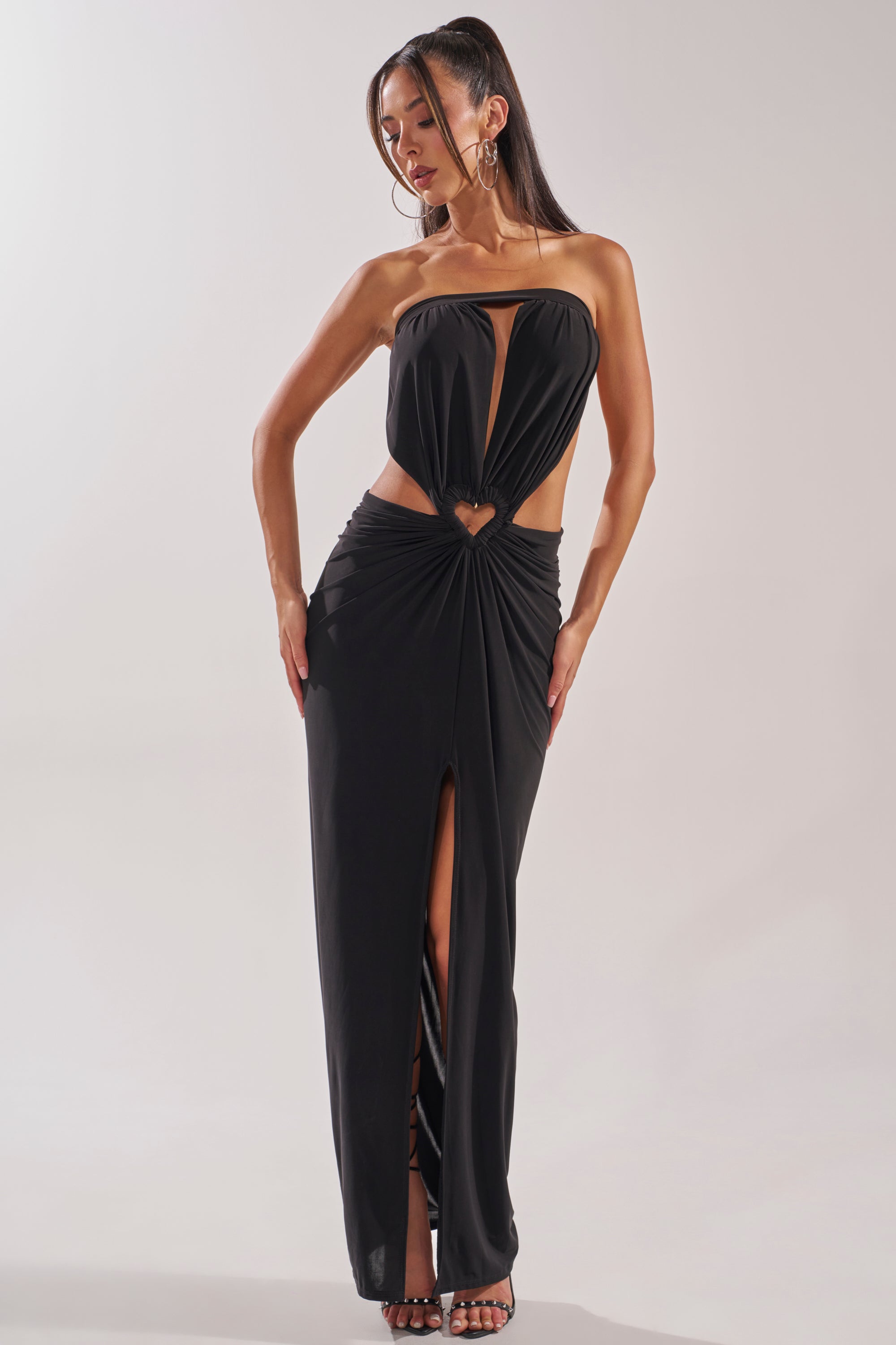 A woman stands hands on hips, looking down while wearing the DONT BREAK MY HEART STRAPLESS MAXI DRESS featuring front cutouts, a keyhole waist detail, and a thigh-high slit against a plain background. 0