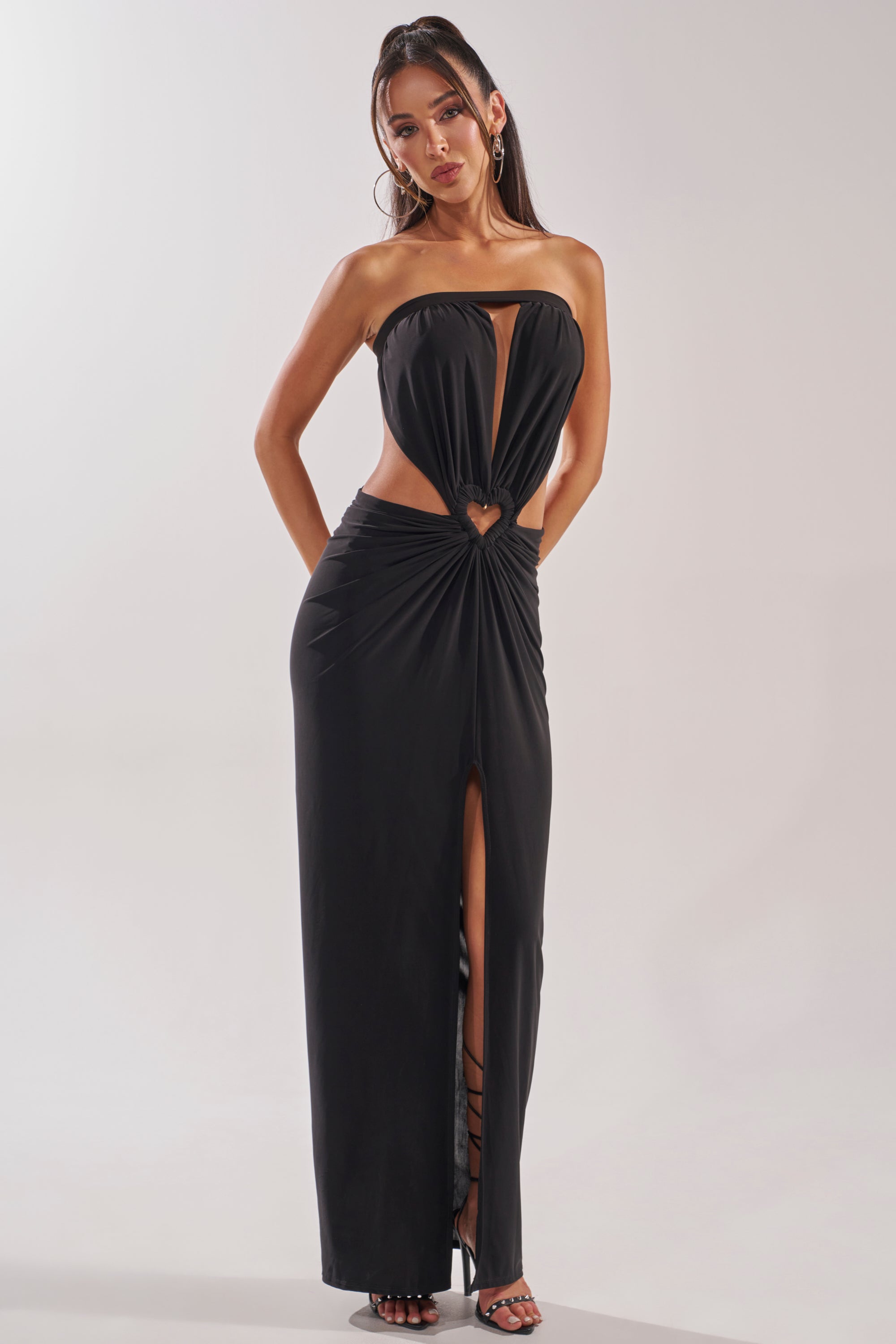 A woman poses against a plain light background wearing the DONT BREAK MY HEART STRAPLESS MAXI DRESS—strapless, floor-length, with front cutouts, heart-shaped waist accent, front slit—and high-heeled sandals. 1