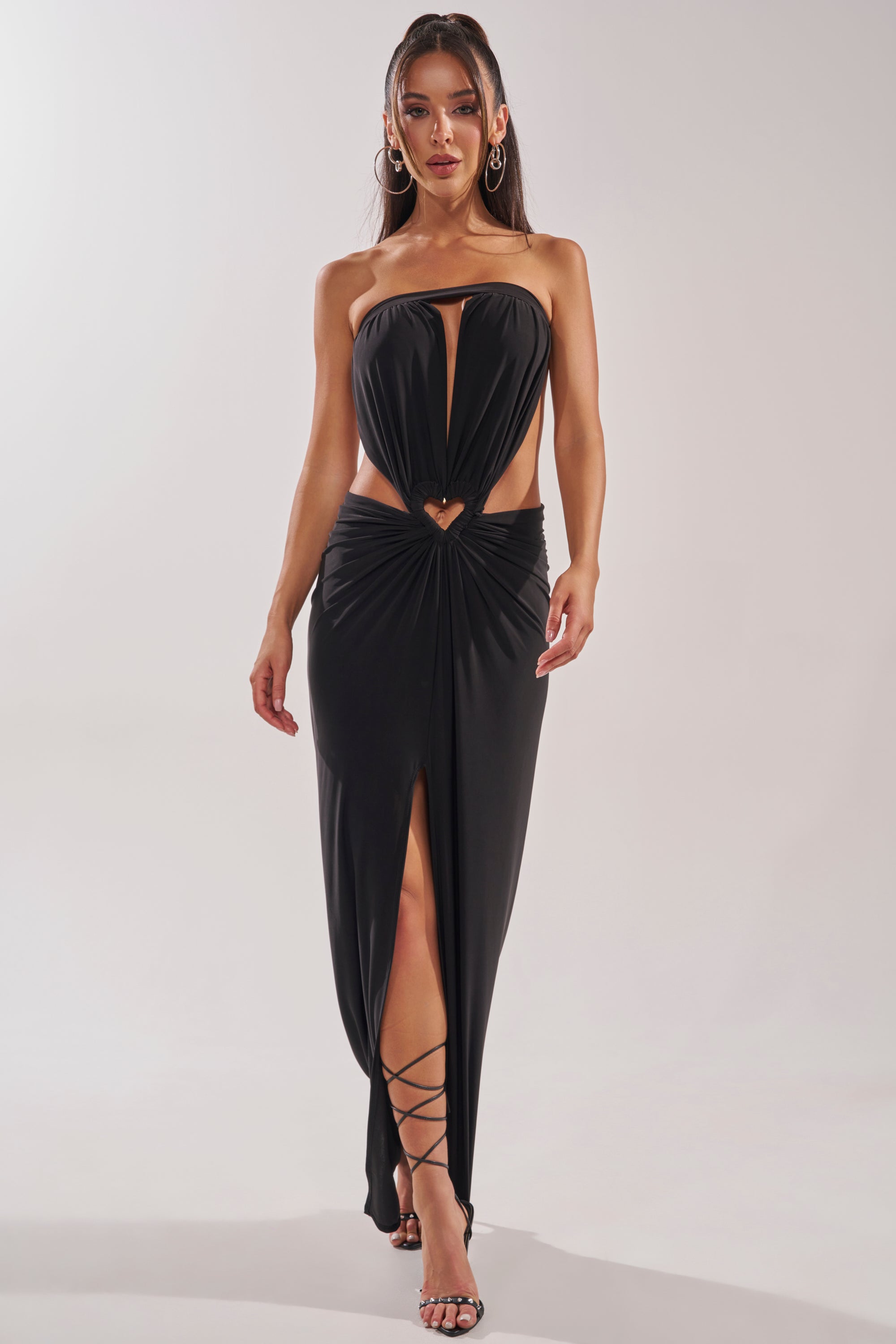 A woman walks confidently toward the camera wearing the DONT BREAK MY HEART STRAPLESS MAXI DRESS, a black formal evening gown with a front keyhole cutout, gathered waist, plunging slit, and strappy high-heeled sandals. 2
