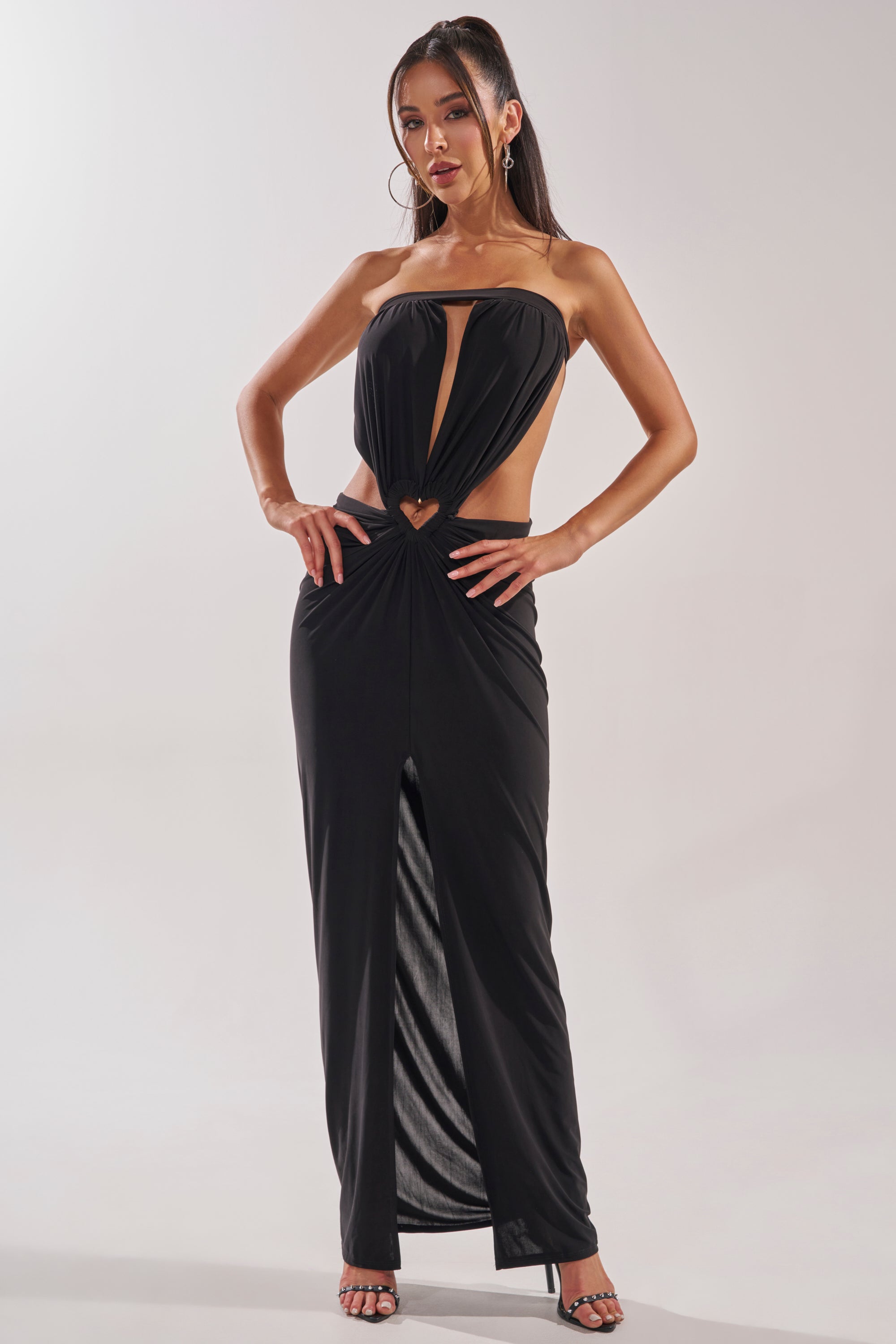 A woman poses confidently in the DONT BREAK MY HEART STRAPLESS MAXI DRESS, featuring a black strapless design, front cutout, and heart detail at the waist. She pairs it with hoop earrings and black heels against a plain light background. 3