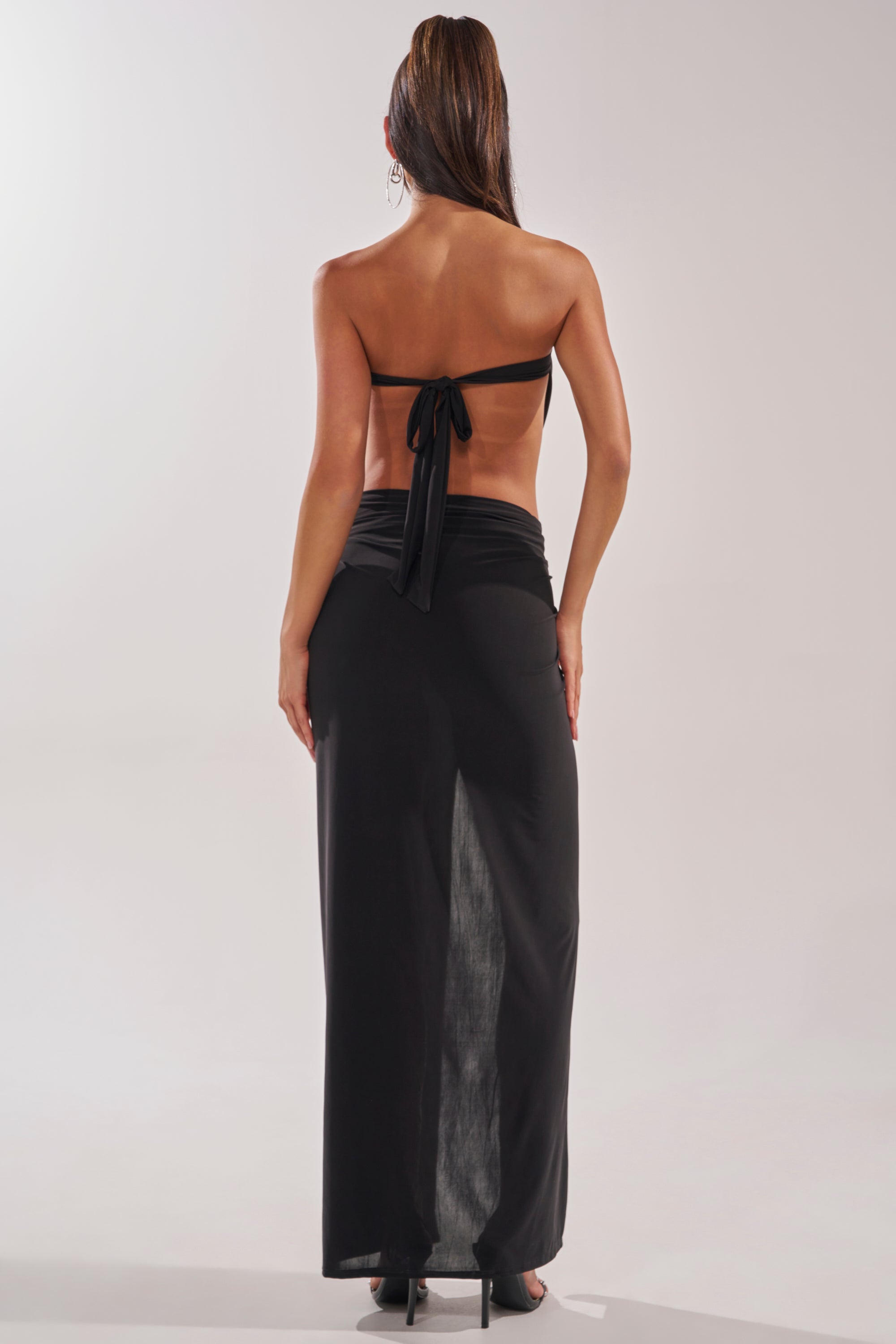 A woman stands facing away, wearing the DONT BREAK MY HEART STRAPLESS MAXI DRESS with a strapless back-tie top and long black skirt. Her hair is in a ponytail, posing against a plain light background. 5