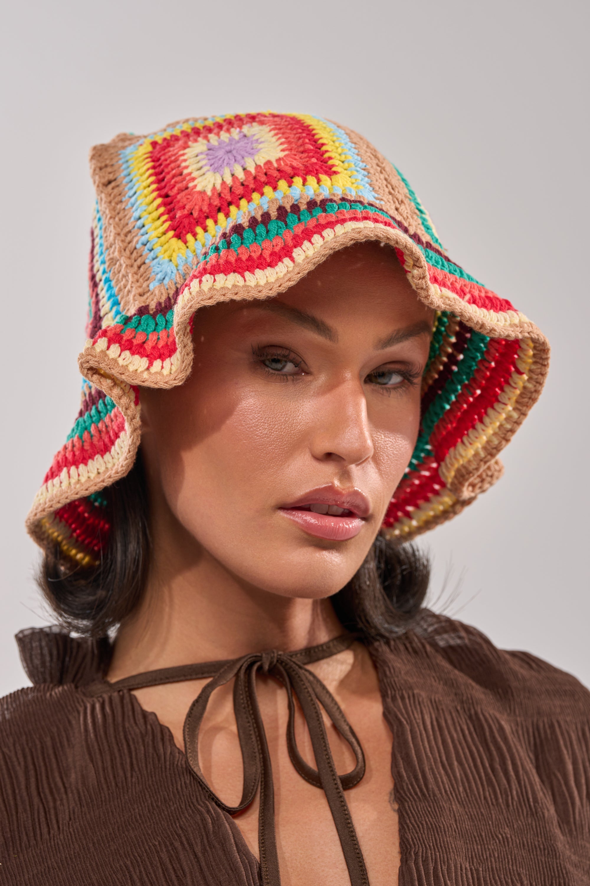 A woman models the NEUTRAL VIBES CROCHET BUCKET HAT, showcasing its colorful granny-square design in red, yellow, blue, and beige against a neutral background. 2