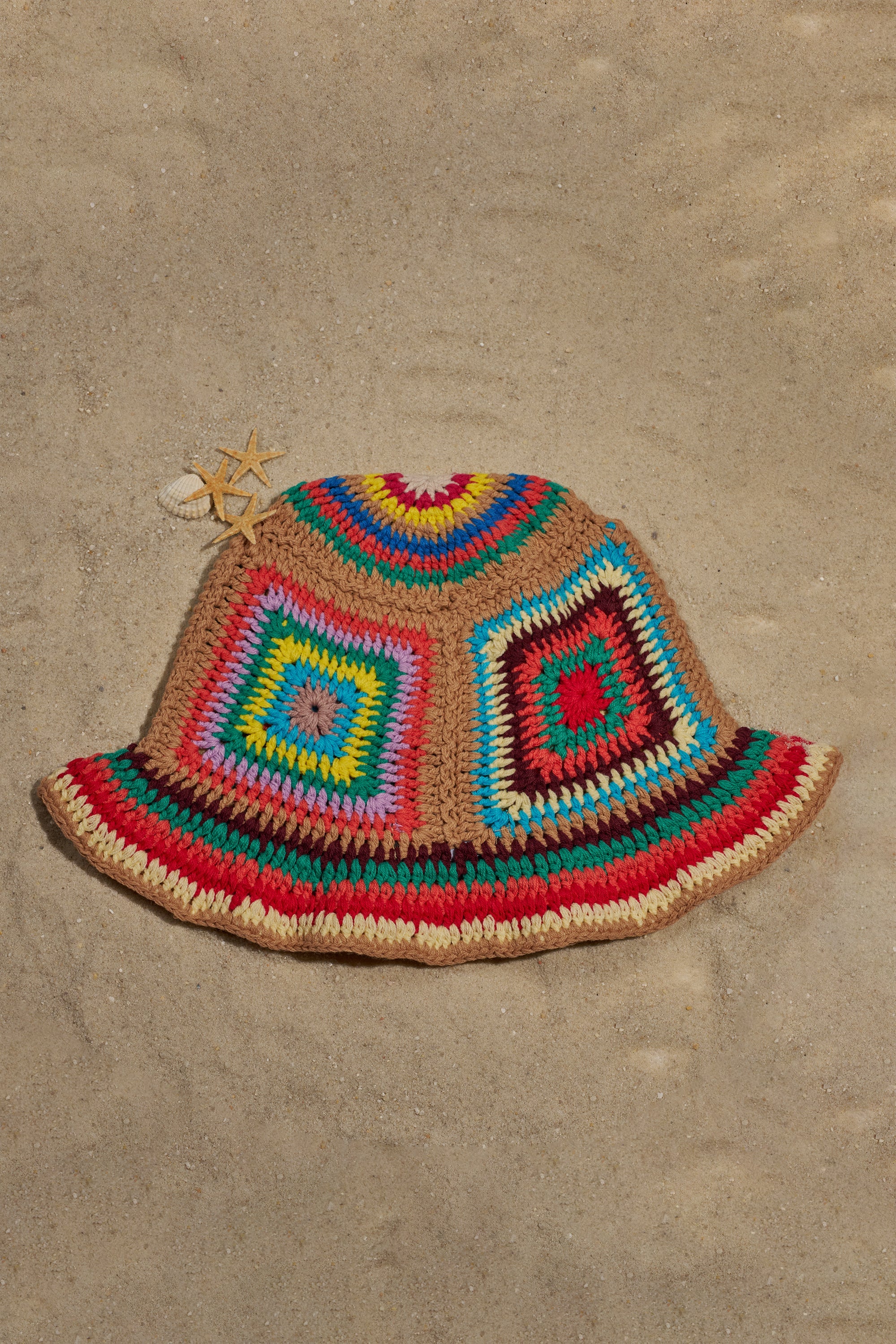 The NEUTRAL VIBES CROCHET BUCKET HAT, featuring a vibrant granny-square crochet design with a shell and starfish accent, is displayed on sandy ground. 1