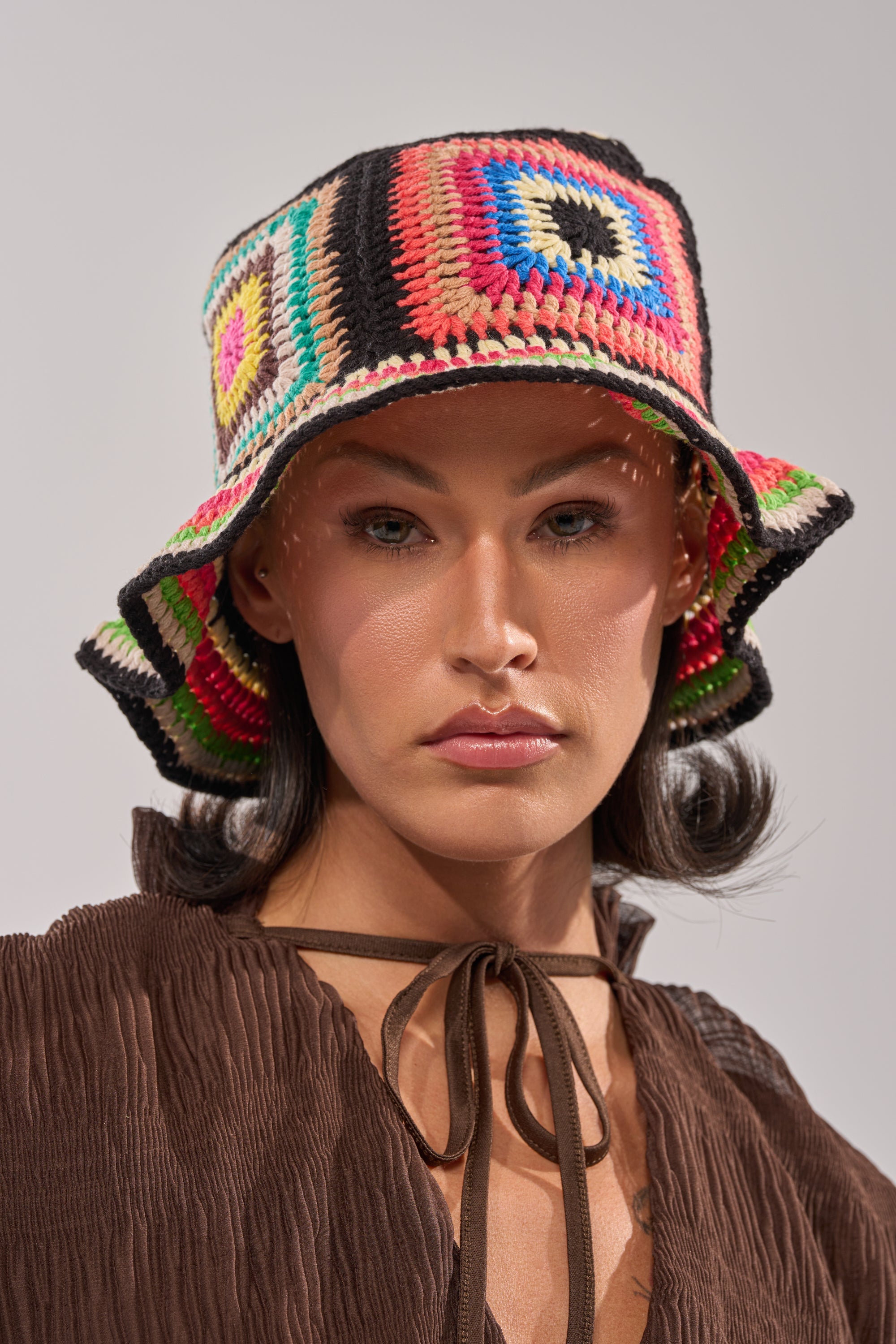 A woman sports the DND CROCHET BUCKET HAT with vibrant geometric crochet patterns, pairing it with a brown textured top and tied neckline, gazing at the camera against a neutral backdrop for a retro-cool look. 2